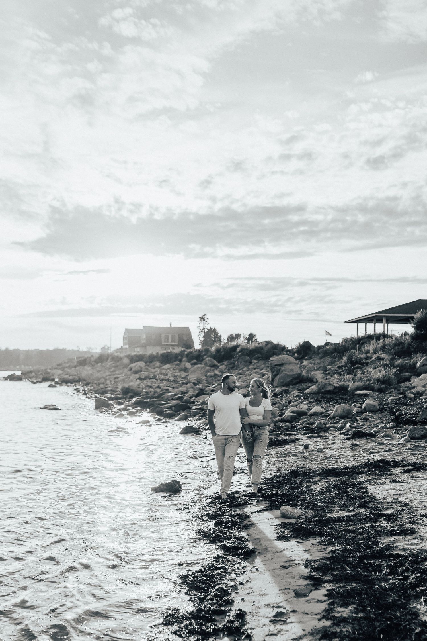 A couple walking along a rocky coastline at sunset with a lighthouse in the distance.