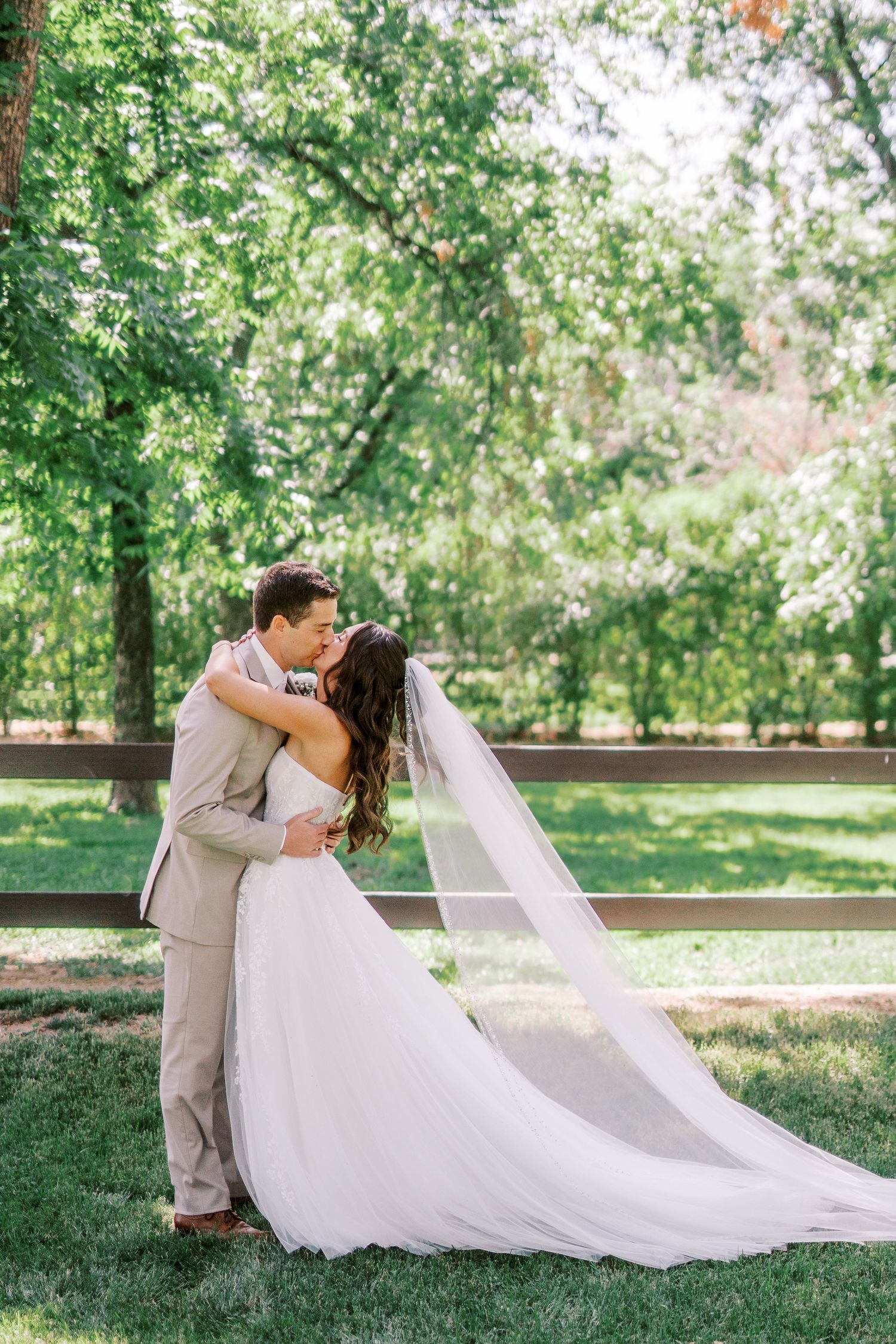 A romantic wedding couple shares a tender moment in a lush green garden setting with a flowing white veil and light beige suit.
