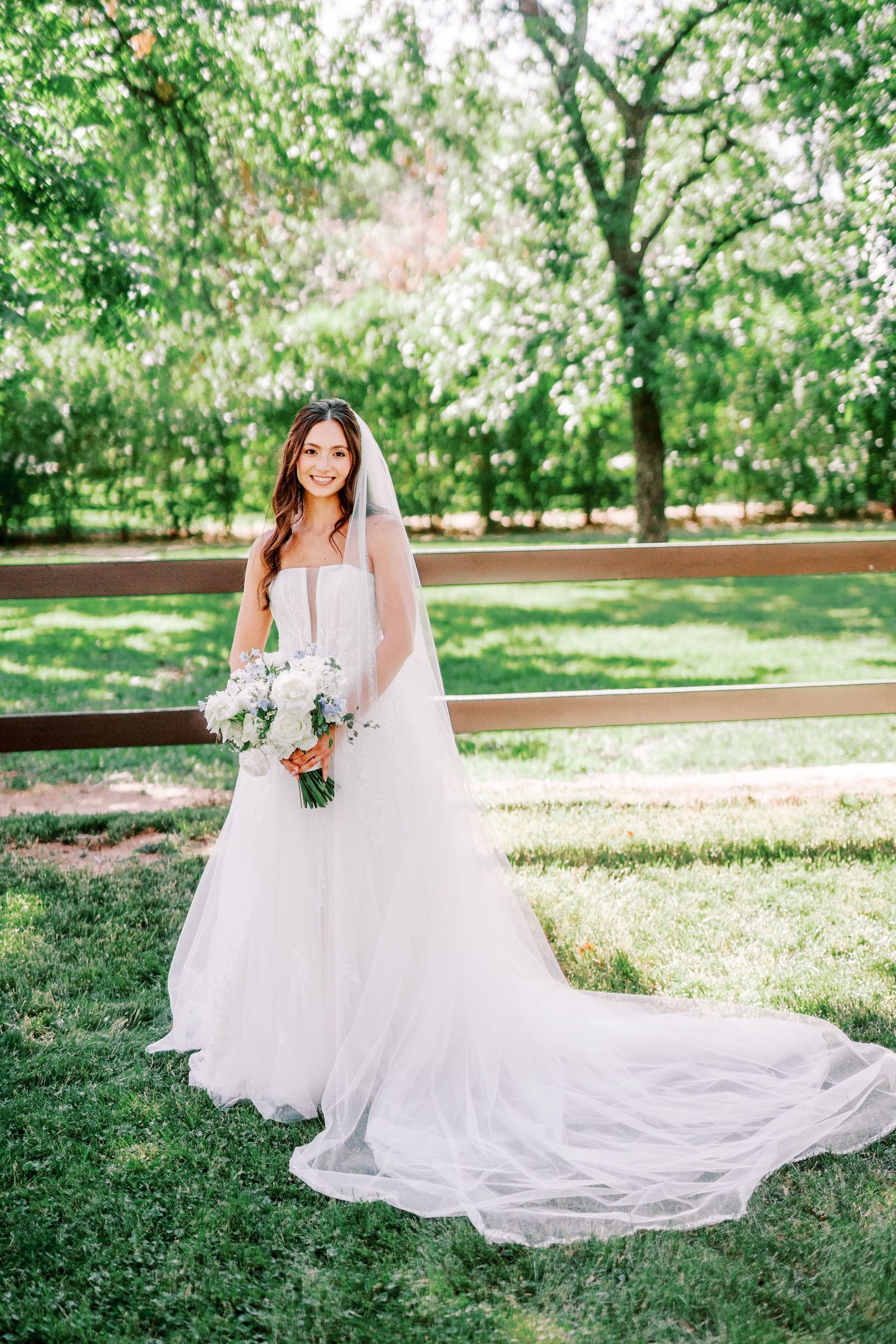 A person in an elegant white wedding gown with a flowing train poses outdoors in a sunlit garden setting with trees.