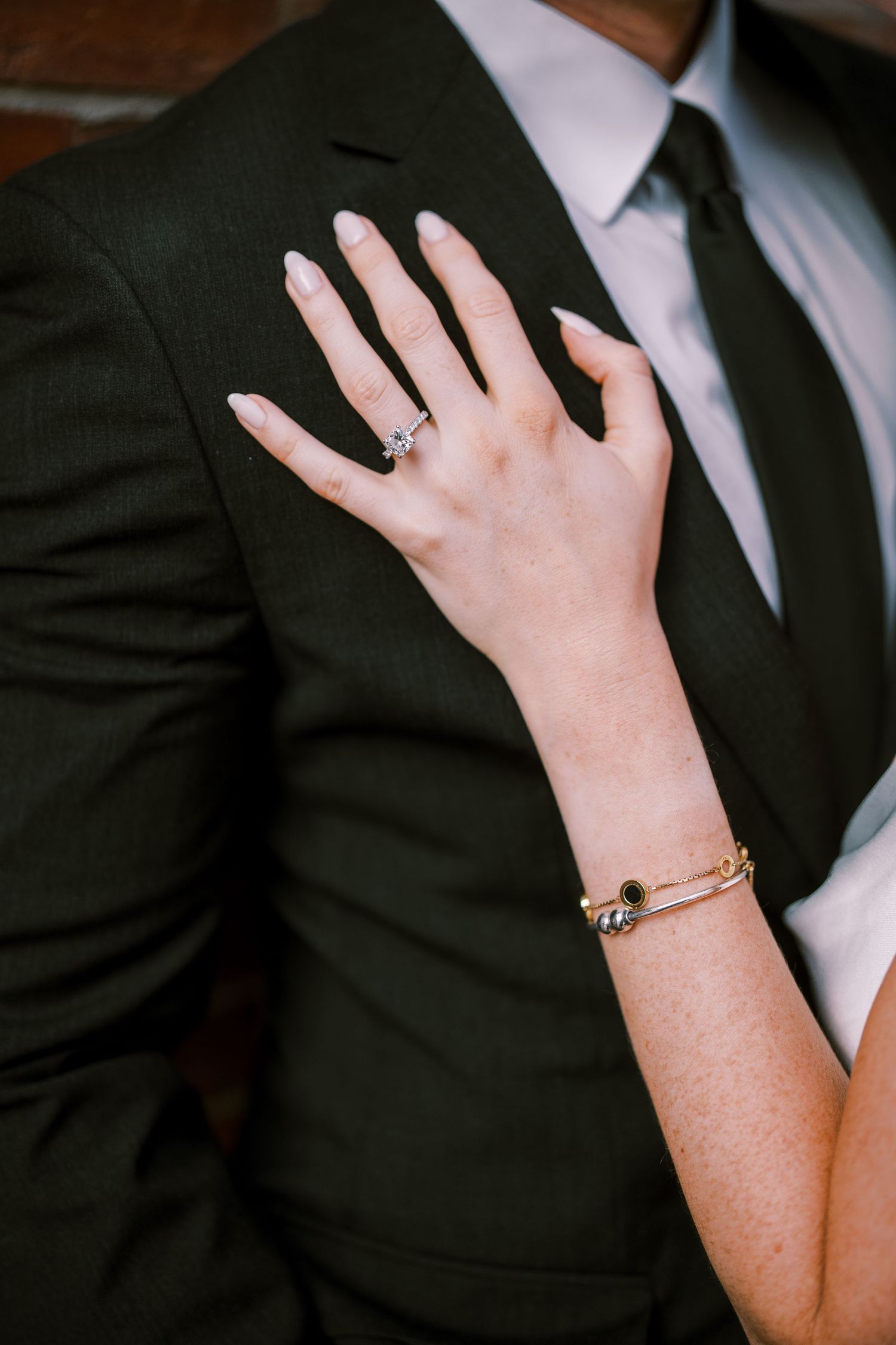 A close-up of a woman's hand with an engagement ring resting on someone wearing a dark suit and tie.