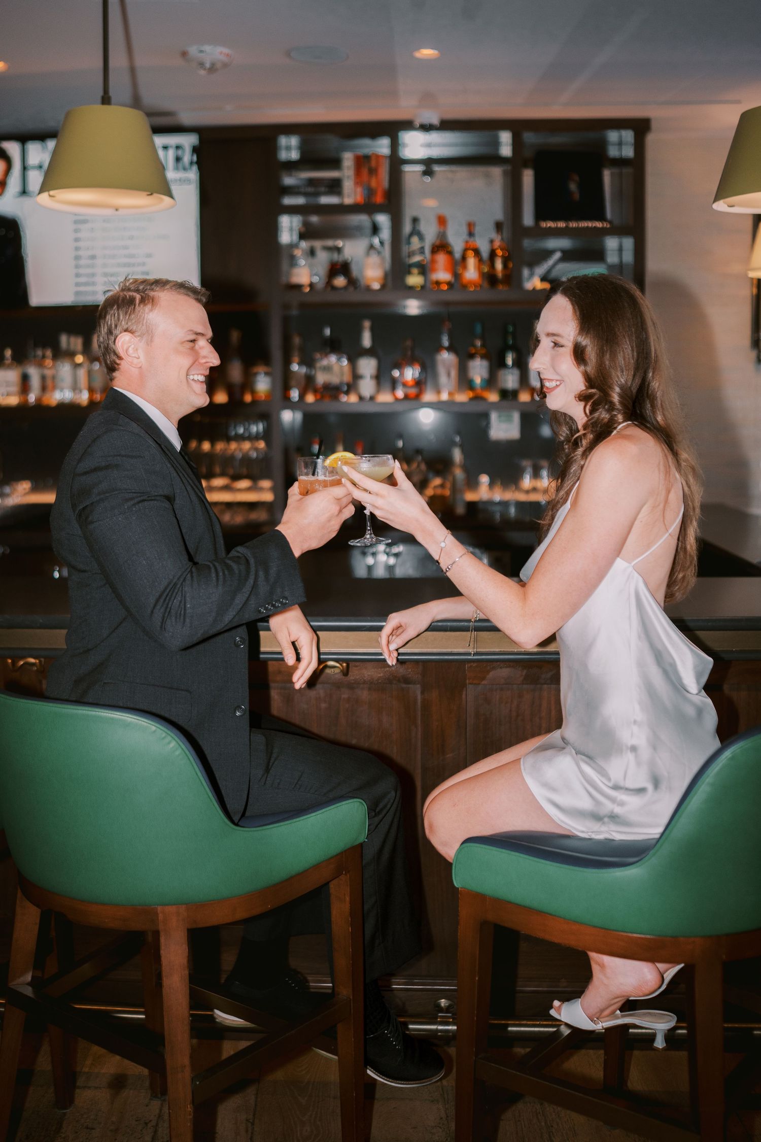 Two people seated at a bar with green stools toast their drinks in a romantic moment.