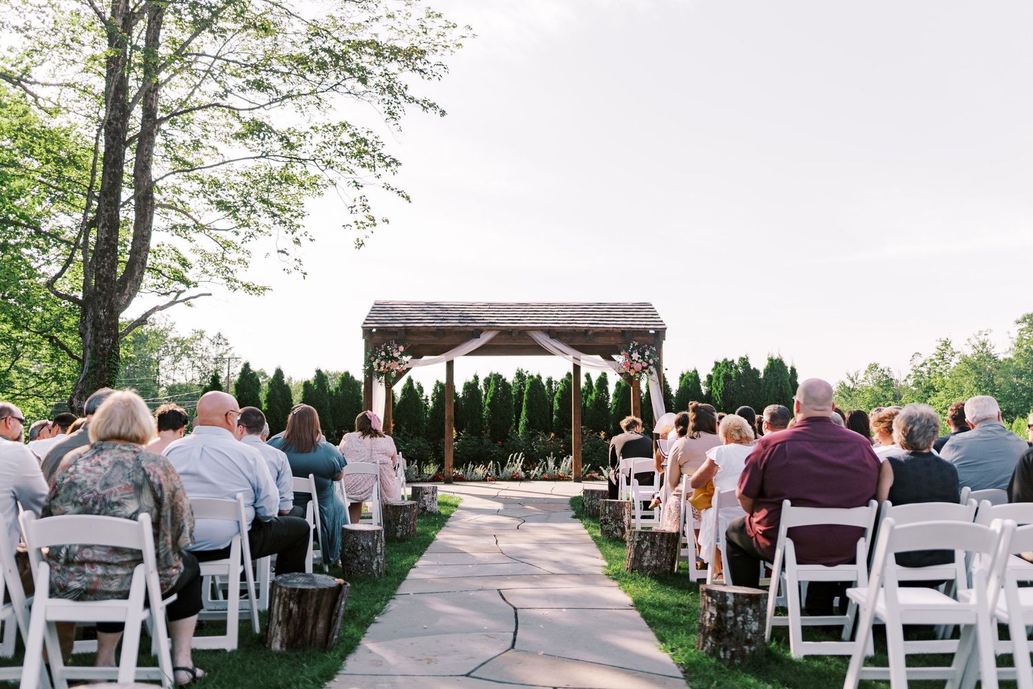Outdoor wedding ceremony setup with white chairs facing a wooden pergola surrounded by greenery.