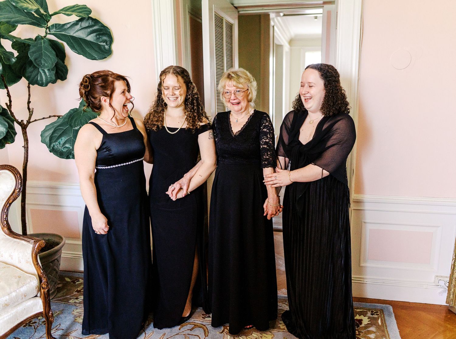 Four women in elegant black dresses share a joyful moment together in an elegant room with pink walls.