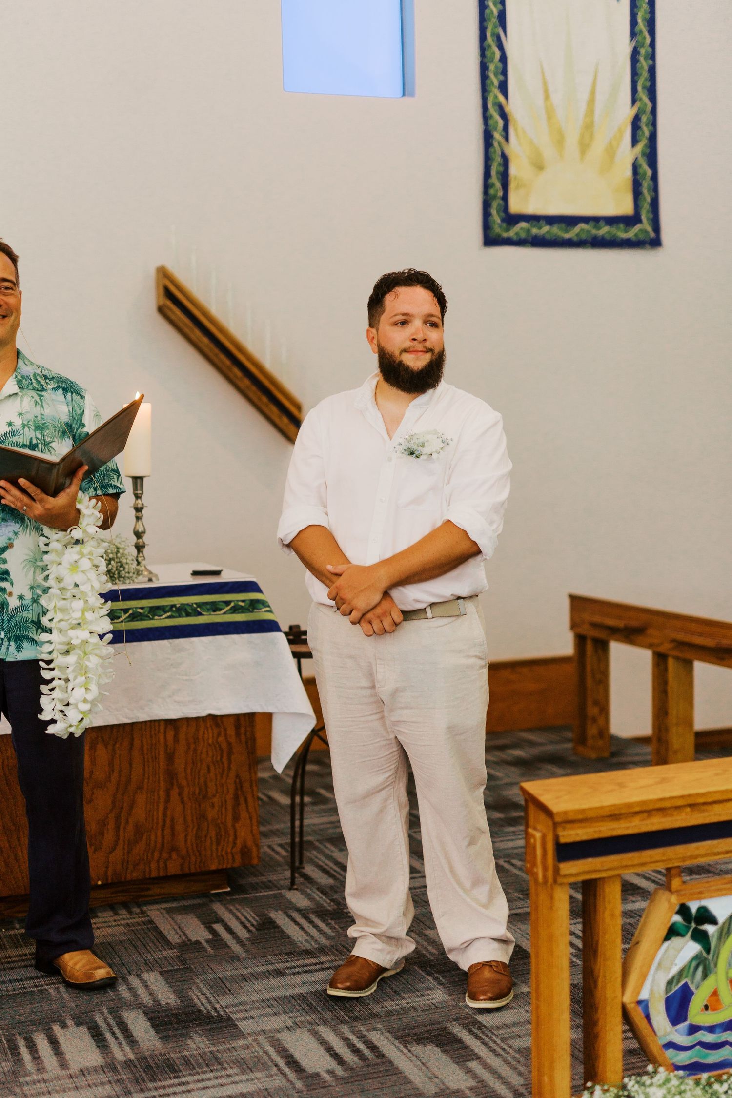 A groom stands in white attire beside an officiant during a wedding ceremony in a church.