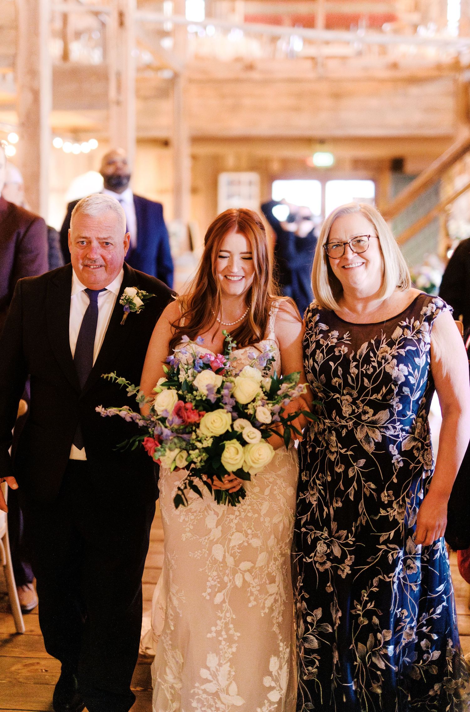 A bride in a lace wedding gown holding a colorful bouquet poses with family at an indoor venue.