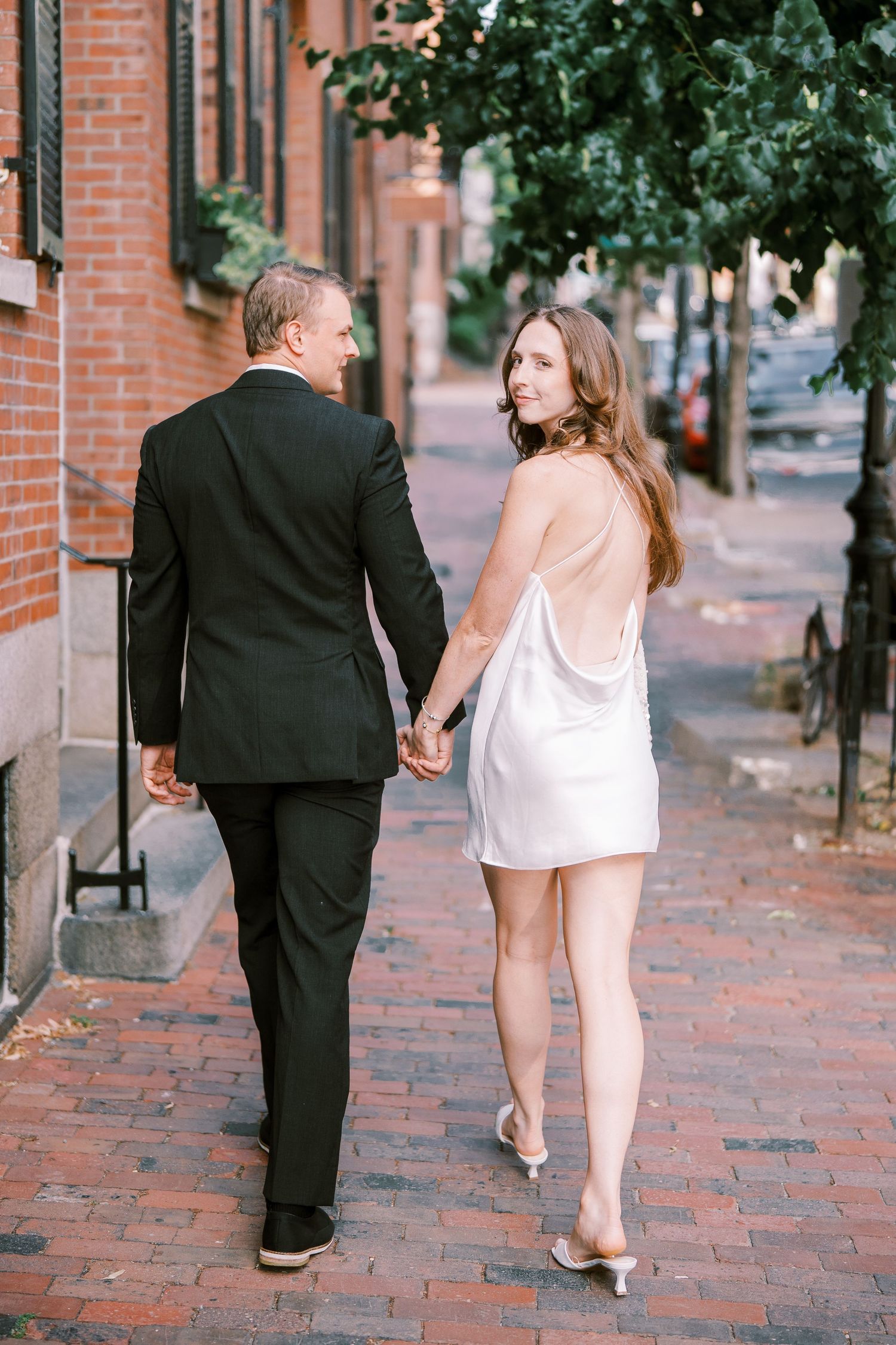 A couple walking hand in hand on a brick sidewalk, with him in a dark suit and her in a white dress.