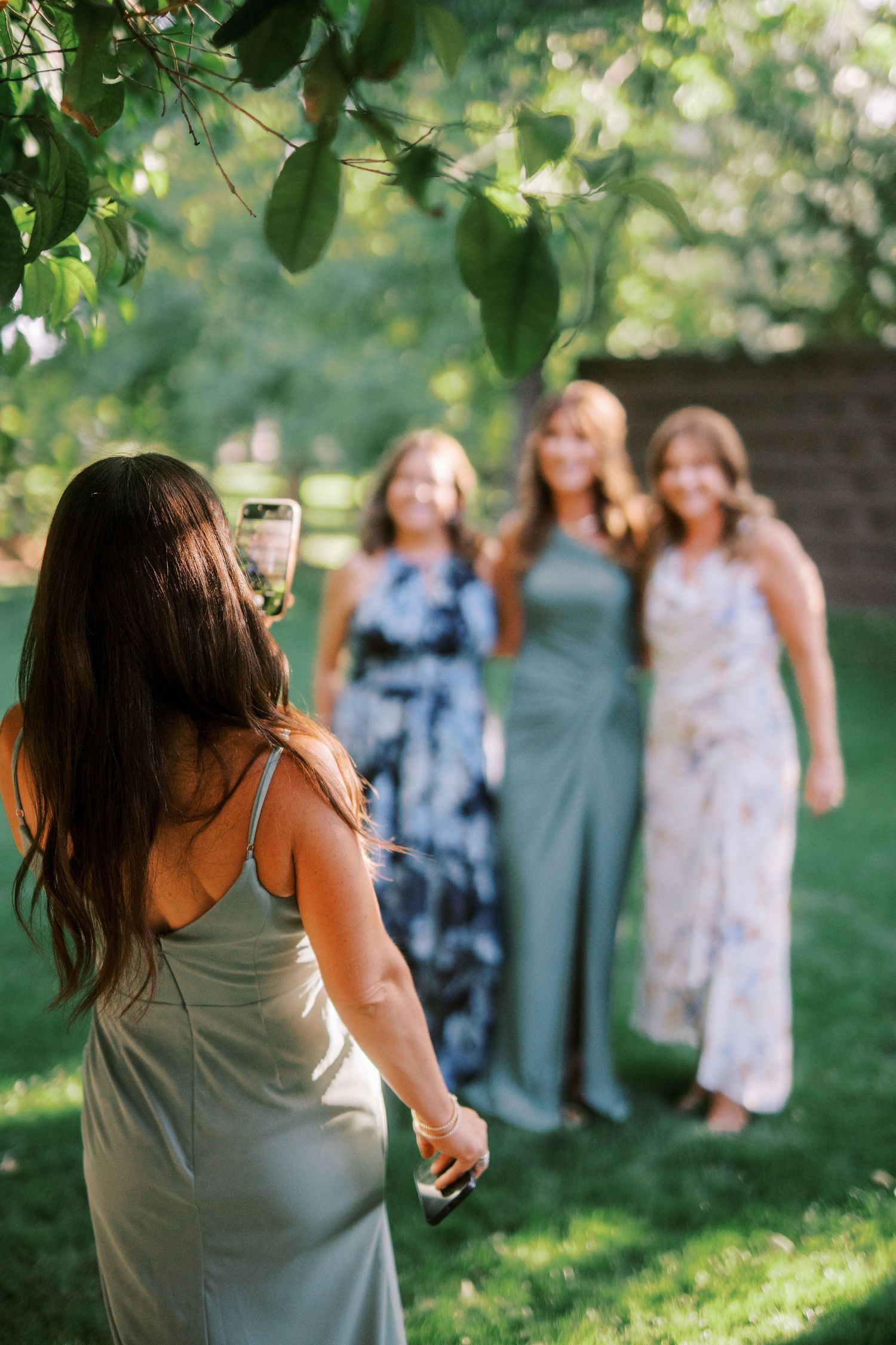 A group in long dresses poses for photos on a sunny lawn under green summer foliage.