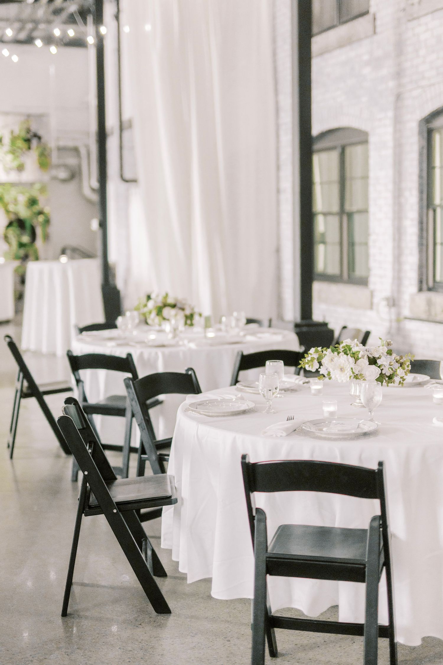 Modern wedding reception setup with white tablecloths and black folding chairs in an industrial space.