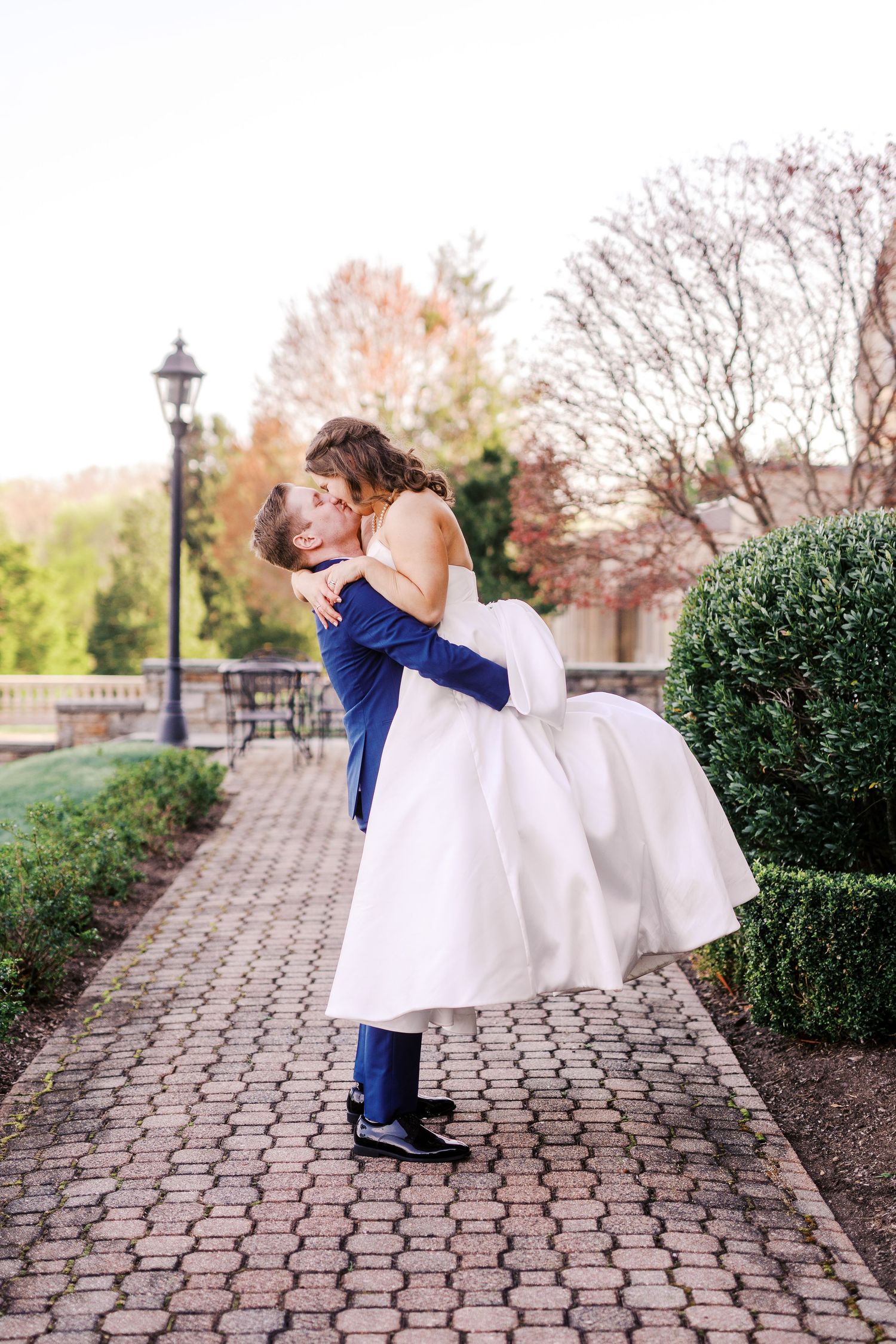 Groom in navy suit lifts bride in white dress during romantic outdoor portrait session.