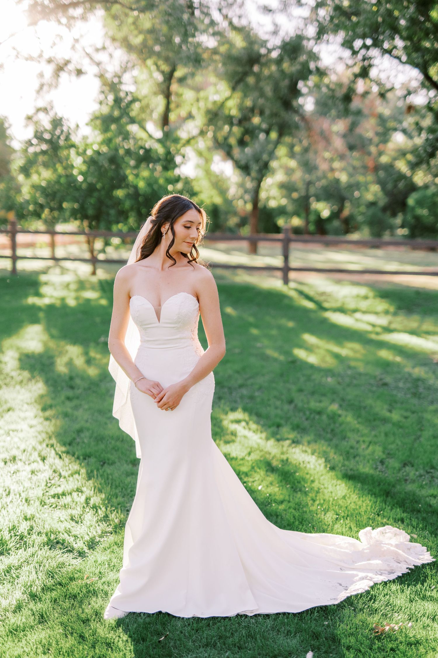 A bride in a flowing white wedding dress sits gracefully on sunlit grass during an outdoor photo session.