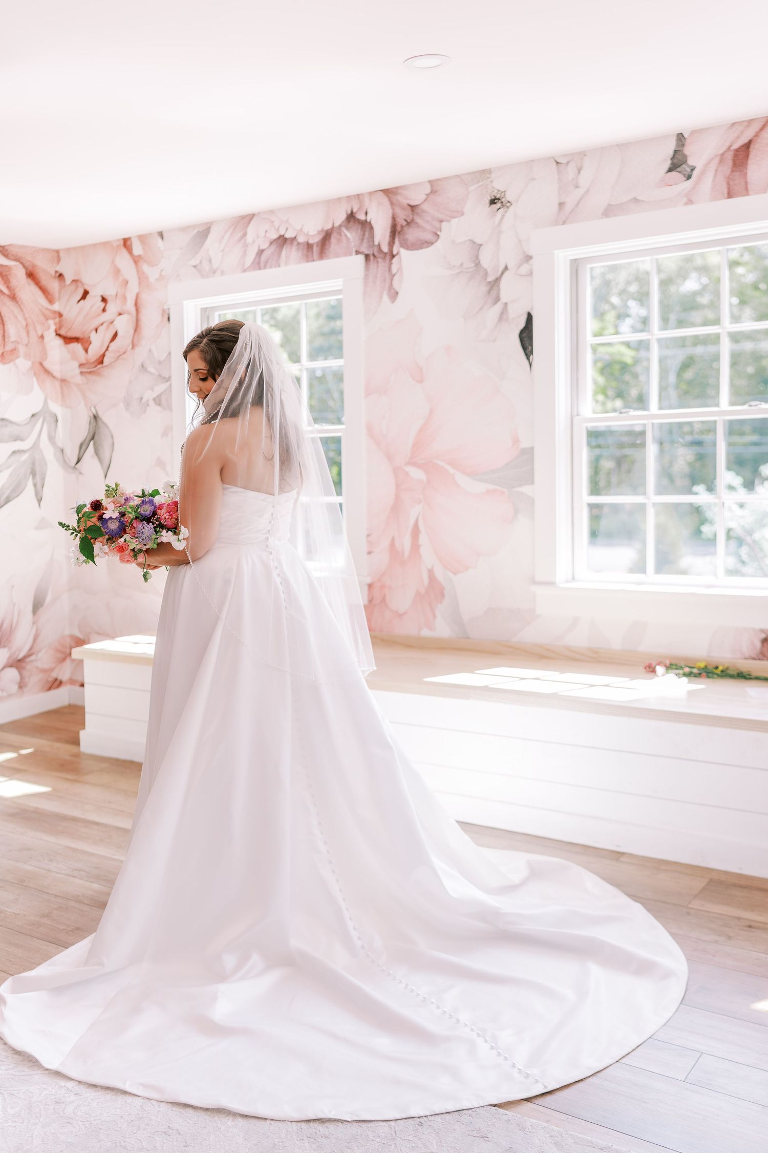 Elegant white wedding dress with dramatic train displayed in a bright room with pink floral wallpaper.