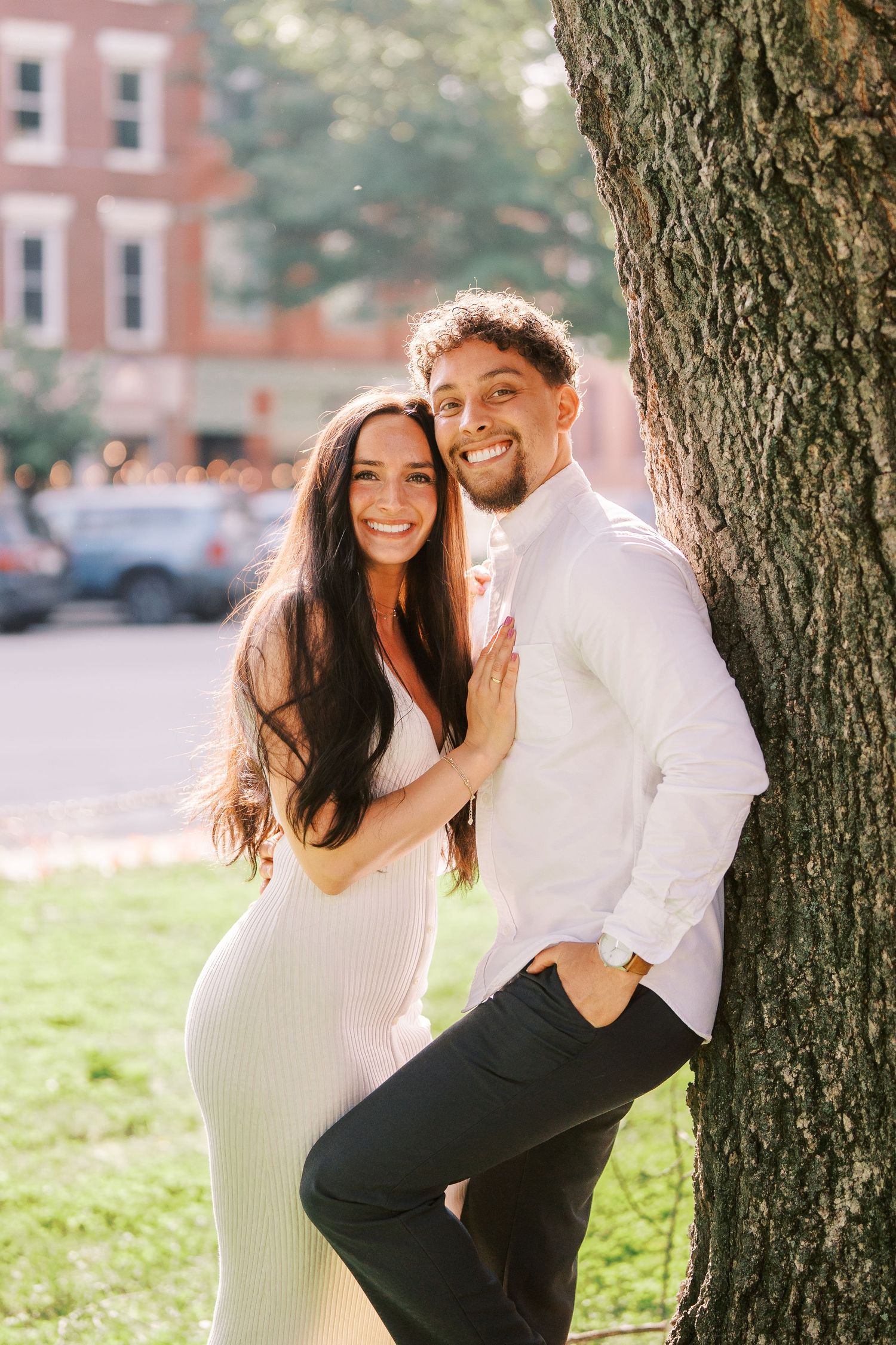 A romantic couple poses together by a large tree trunk in an urban setting.