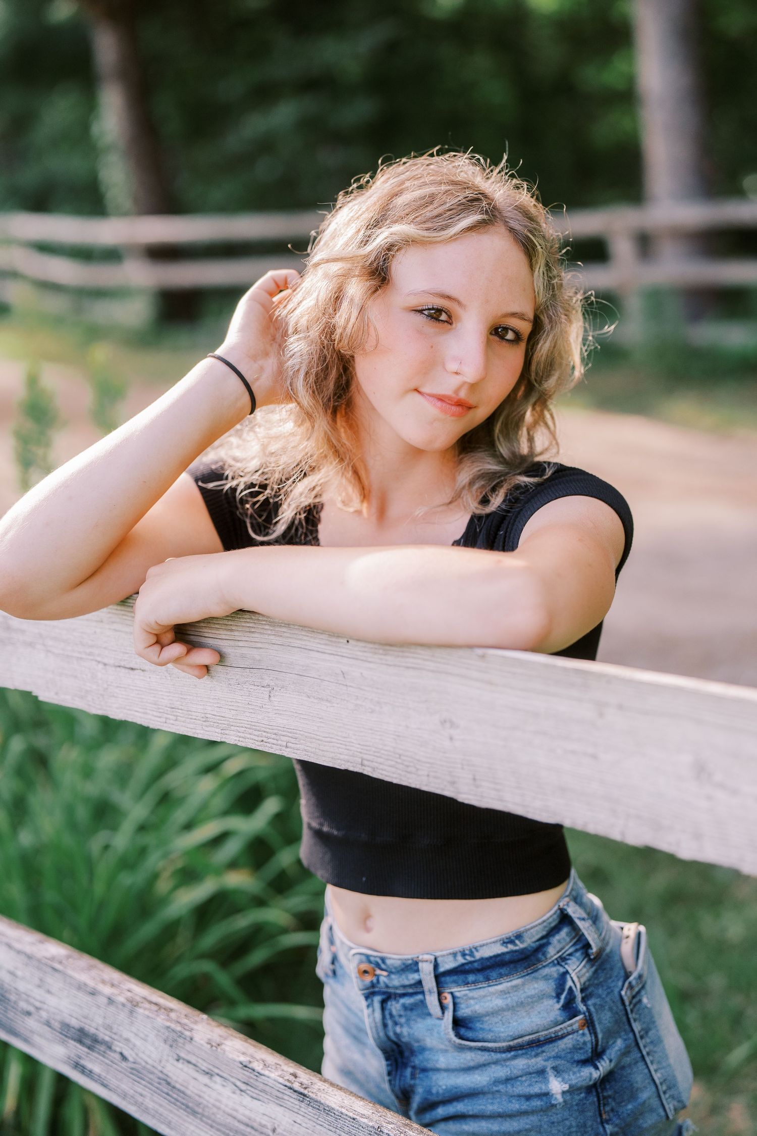 Casual summer portrait of someone in a black crop top and jeans leaning on a wooden fence in a natural outdoor setting.