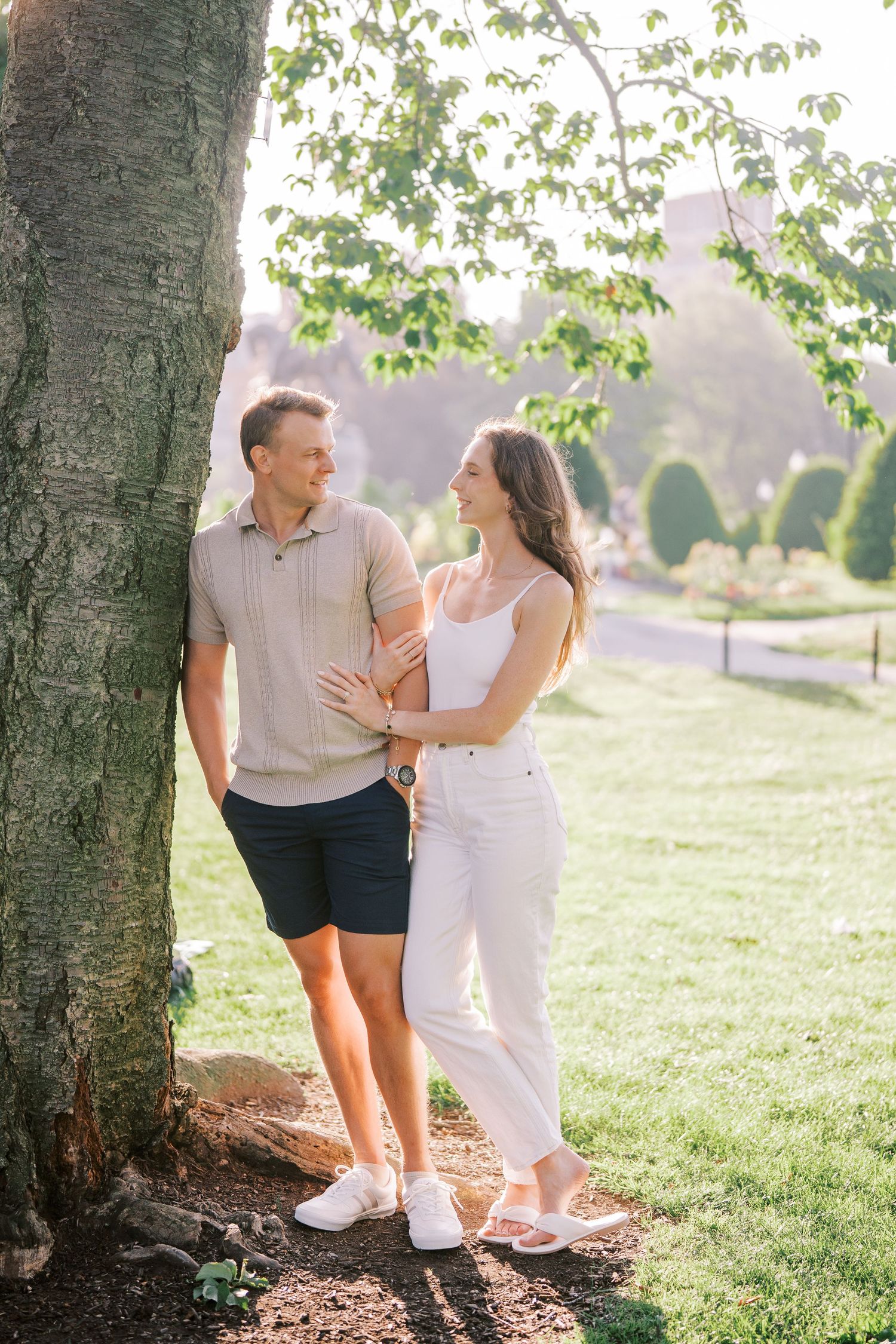 A couple shares a tender moment while leaning against a tree trunk in a sunny park setting during golden hour.