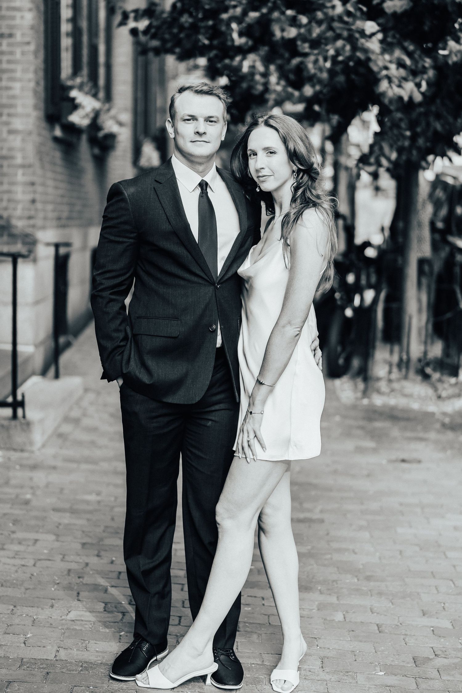 Black and white photo of a couple in formal attire embracing on a brick sidewalk in an urban setting.