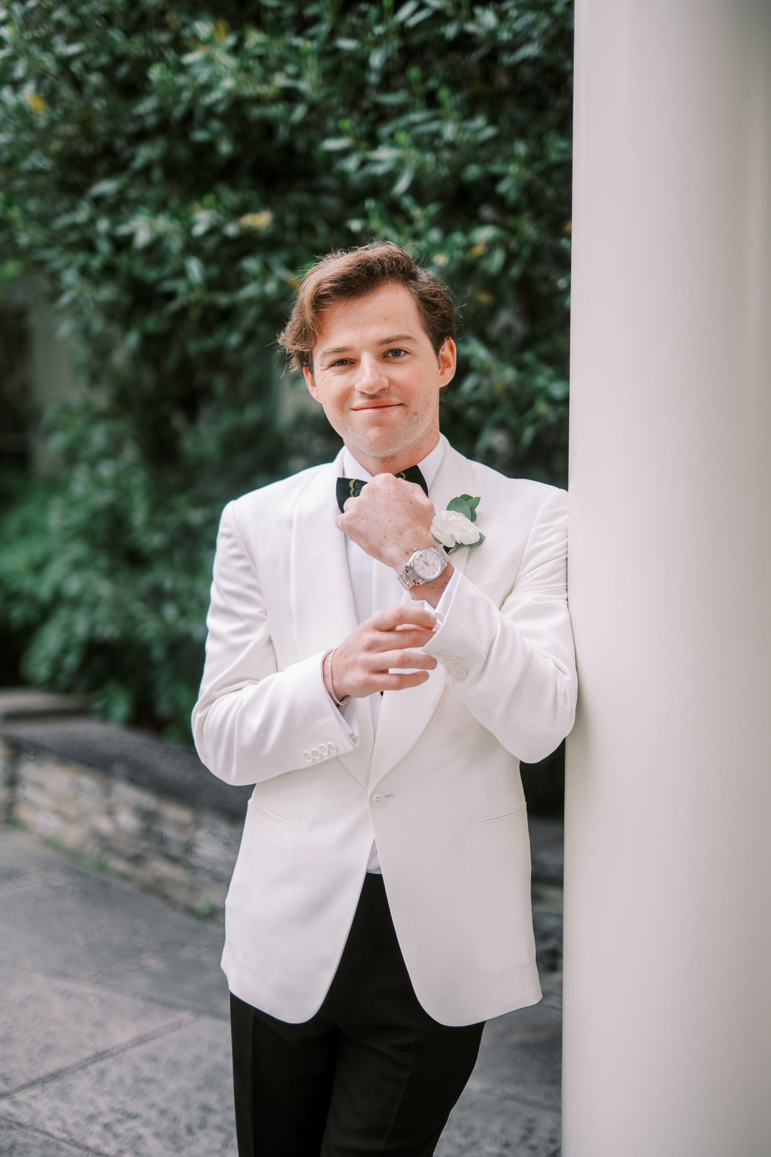 A person in a white dinner jacket and bow tie holding a glass in front of lush greenery at a formal event.