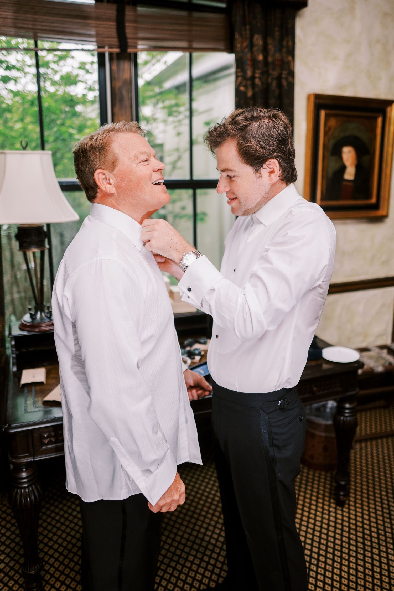 Two people in white dress shirts stand together preparing formal attire in an elegant interior setting.