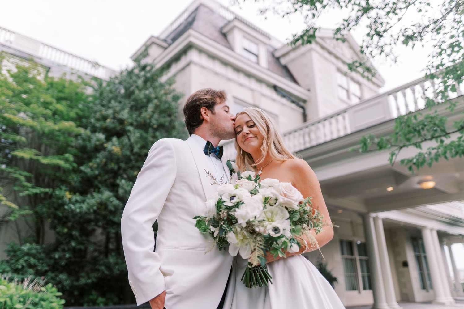 A romantic wedding couple shares joyful moments in front of an elegant white mansion during their outdoor celebration.