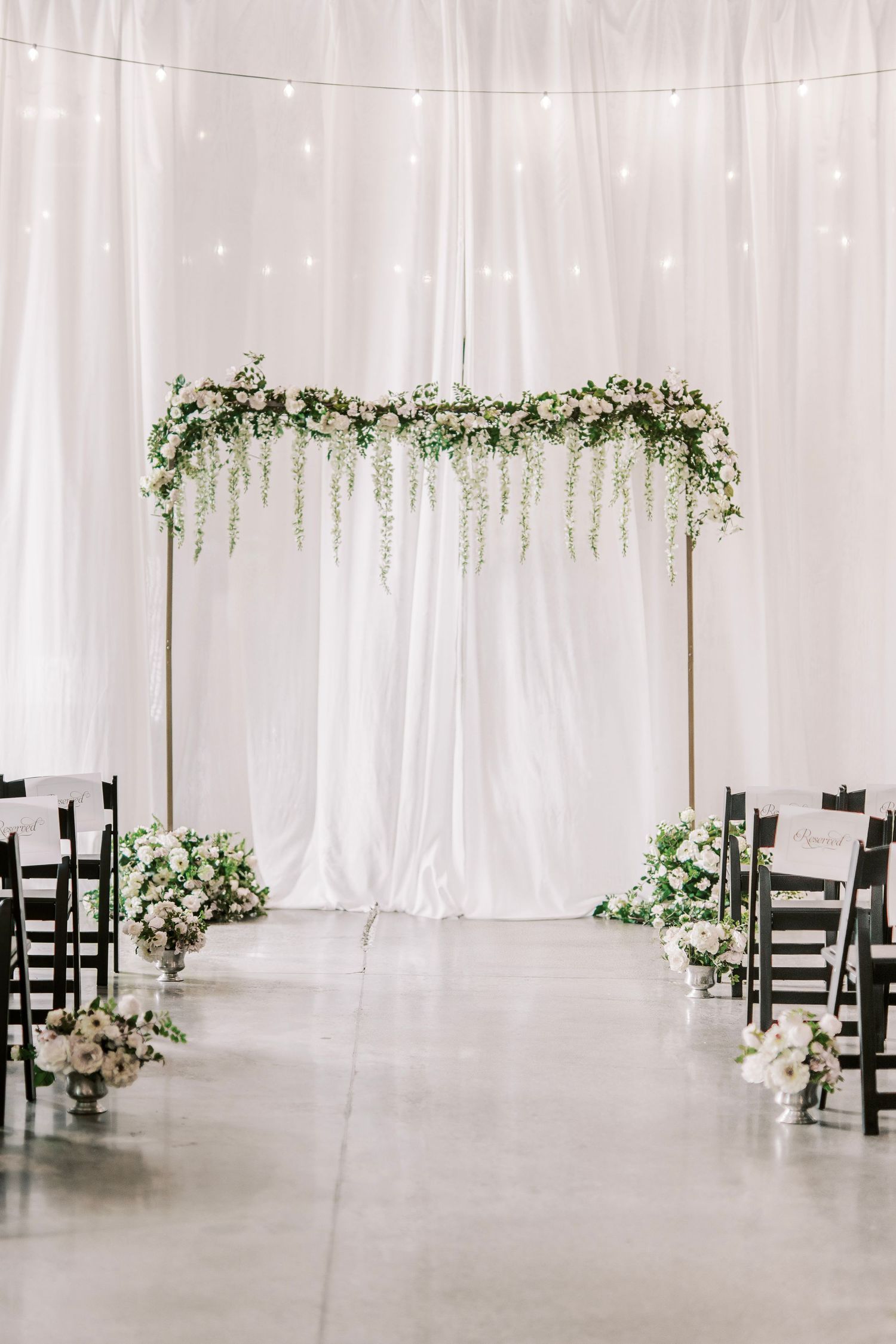 Elegant wedding ceremony setup with white drapes and floral arch decorations in an indoor venue.