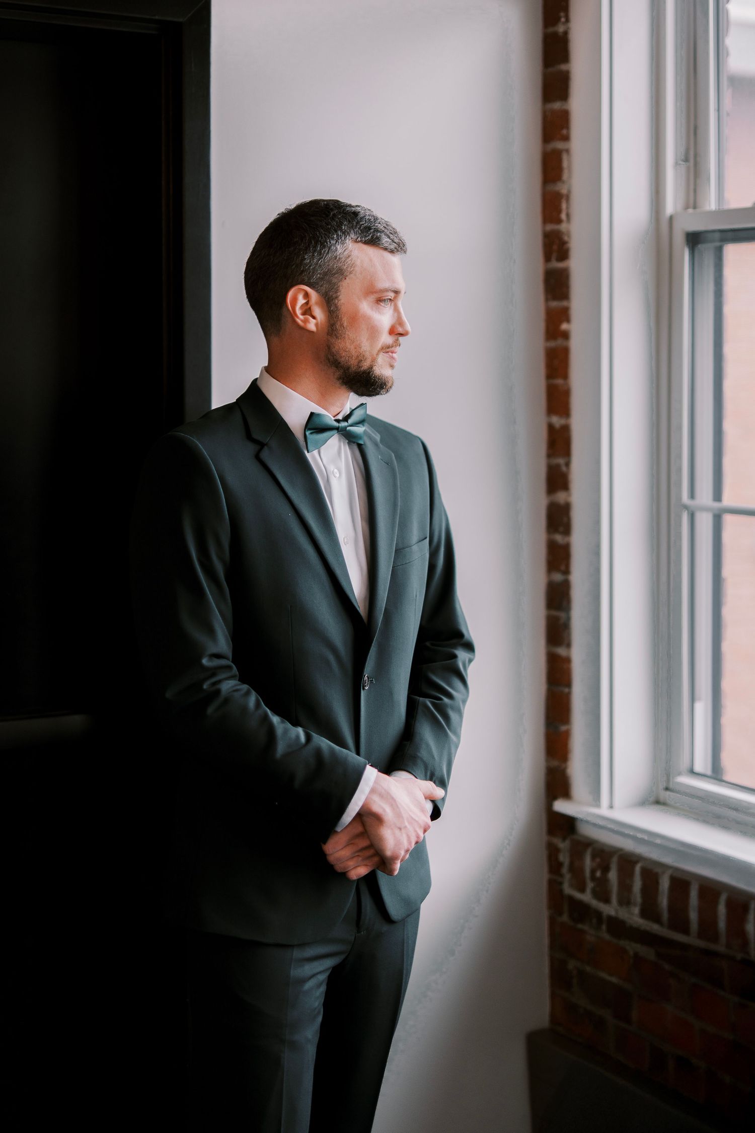 A person in a dark formal suit and bow tie stands contemplatively by a window with exposed brick walls.
