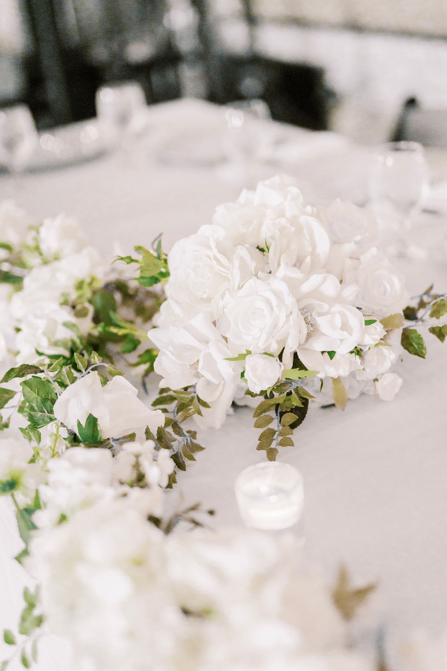 Elegant white floral centerpiece with roses and greenery arranged on a light gray tablecloth at a wedding reception.