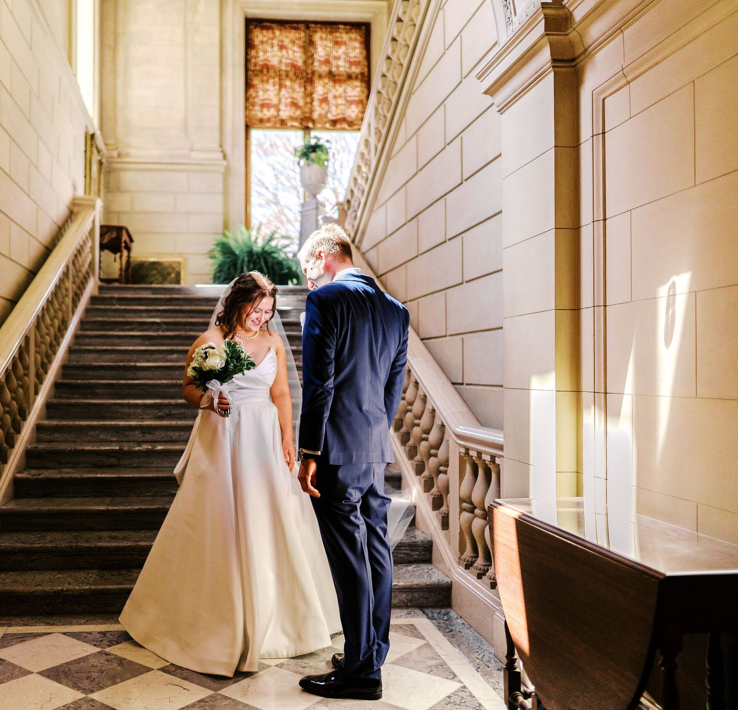 Couple in formal attire stand together on an elegant marble staircase with natural light streaming through windows.