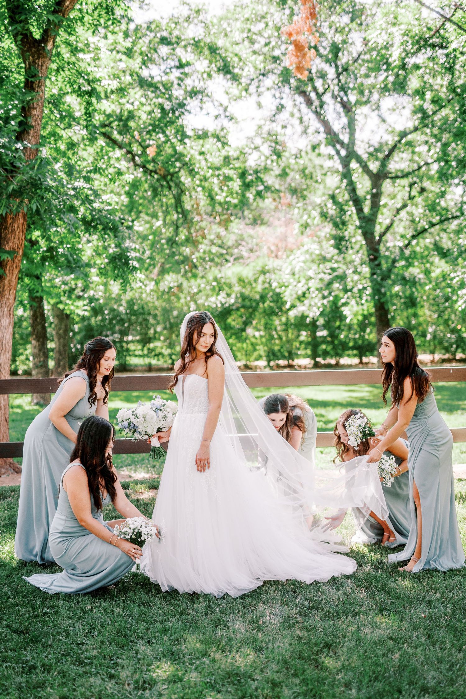 Bridesmaids in light blue dresses help adjust the brides flowing white gown on a sunny day in a green park setting.