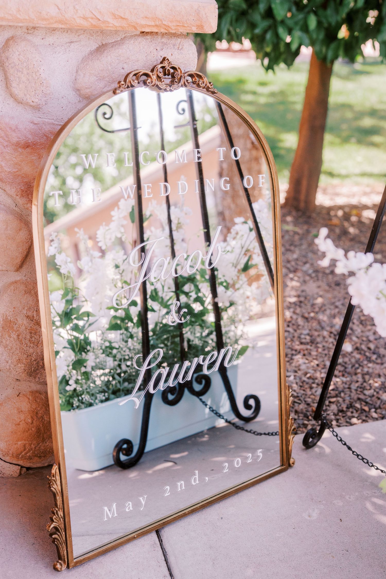 Vintage mirror with elegant scrollwork frame displays white wedding text and delicate white floral arrangements.