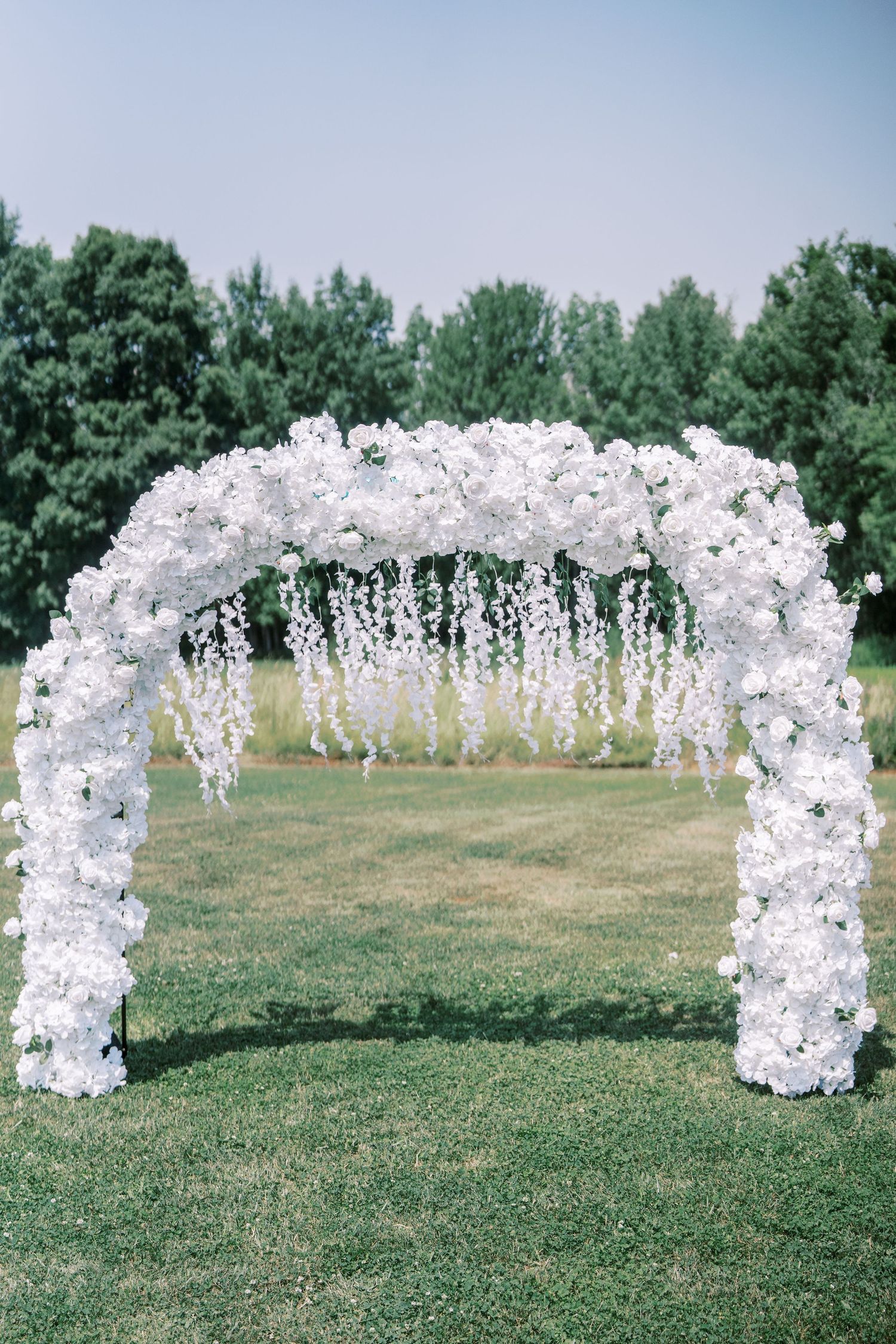 Curved white floral wedding arch frames a green outdoor ceremony space in an elegant garden setting.
