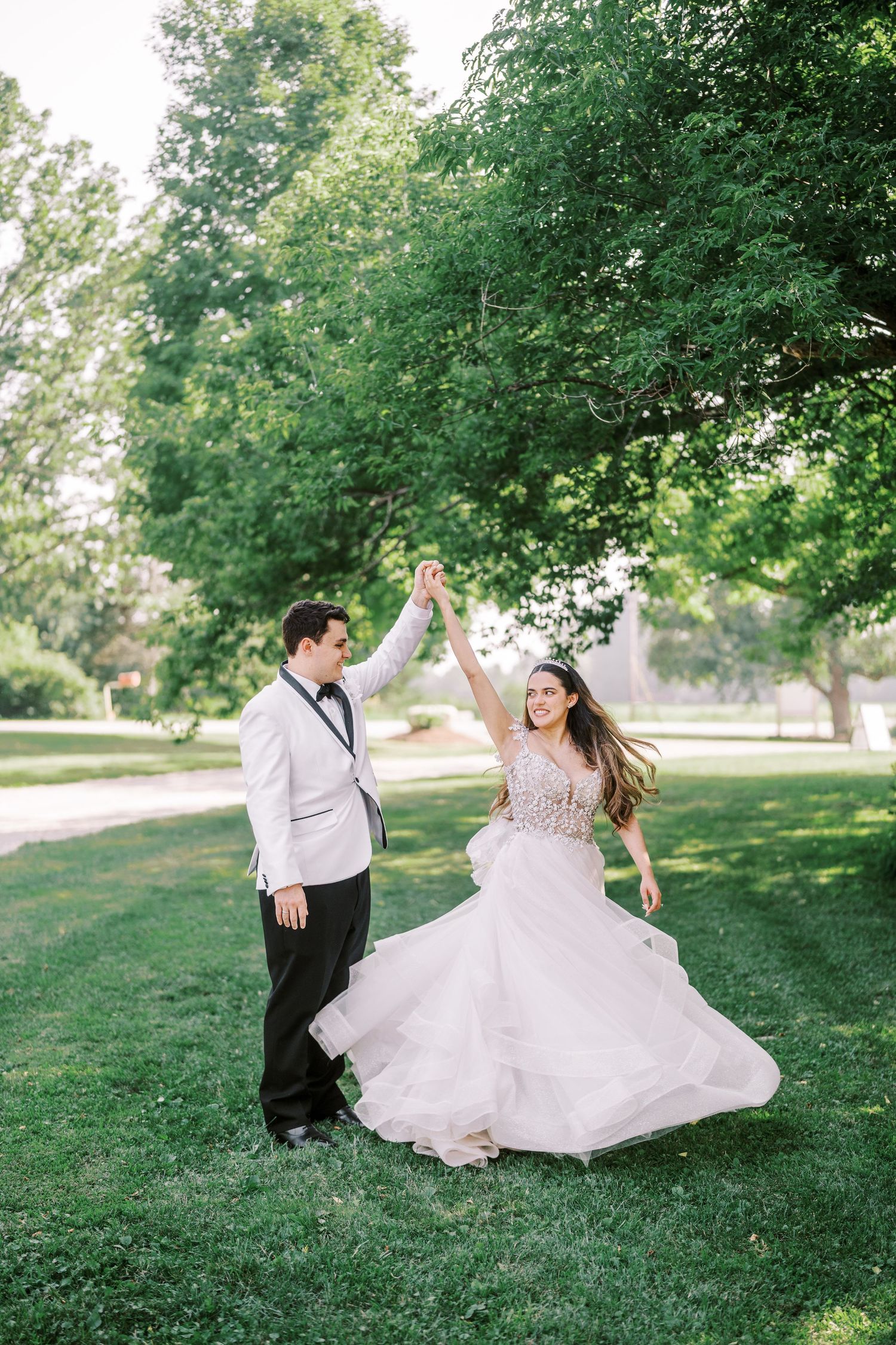 A couple dances together on a lush green lawn with tall trees in the background, her flowing blush gown twirling gracefully.