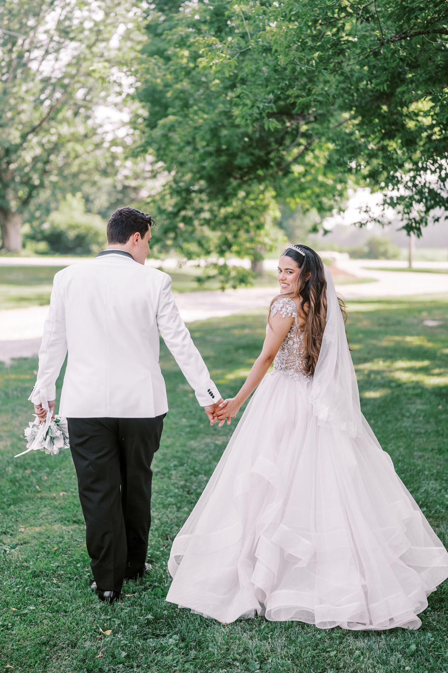 A newlywed couple walks hand in hand on grass under trees, with the bride wearing a flowing white gown and veil.
