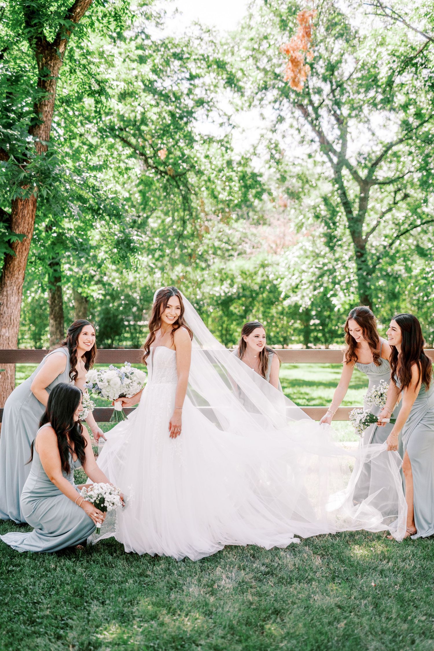A wedding party in light gray dresses helps arrange a flowing white bridal gown and veil on a sunny lawn with trees.