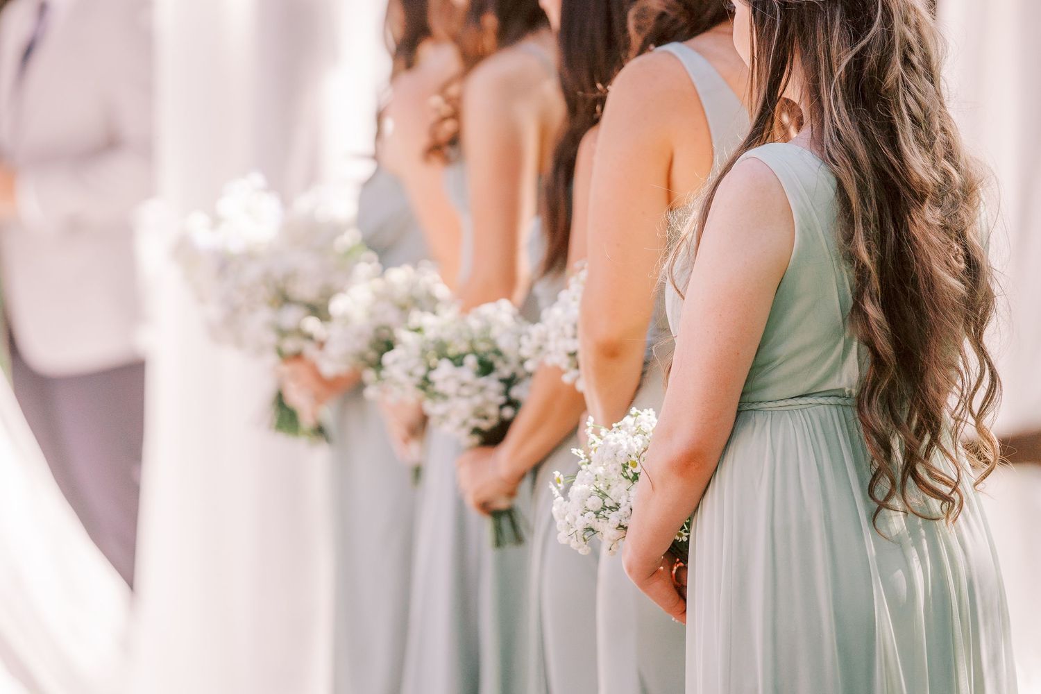 Bridesmaids in soft sage green dresses line up with bouquets showing details of their flowing gowns at a wedding.
