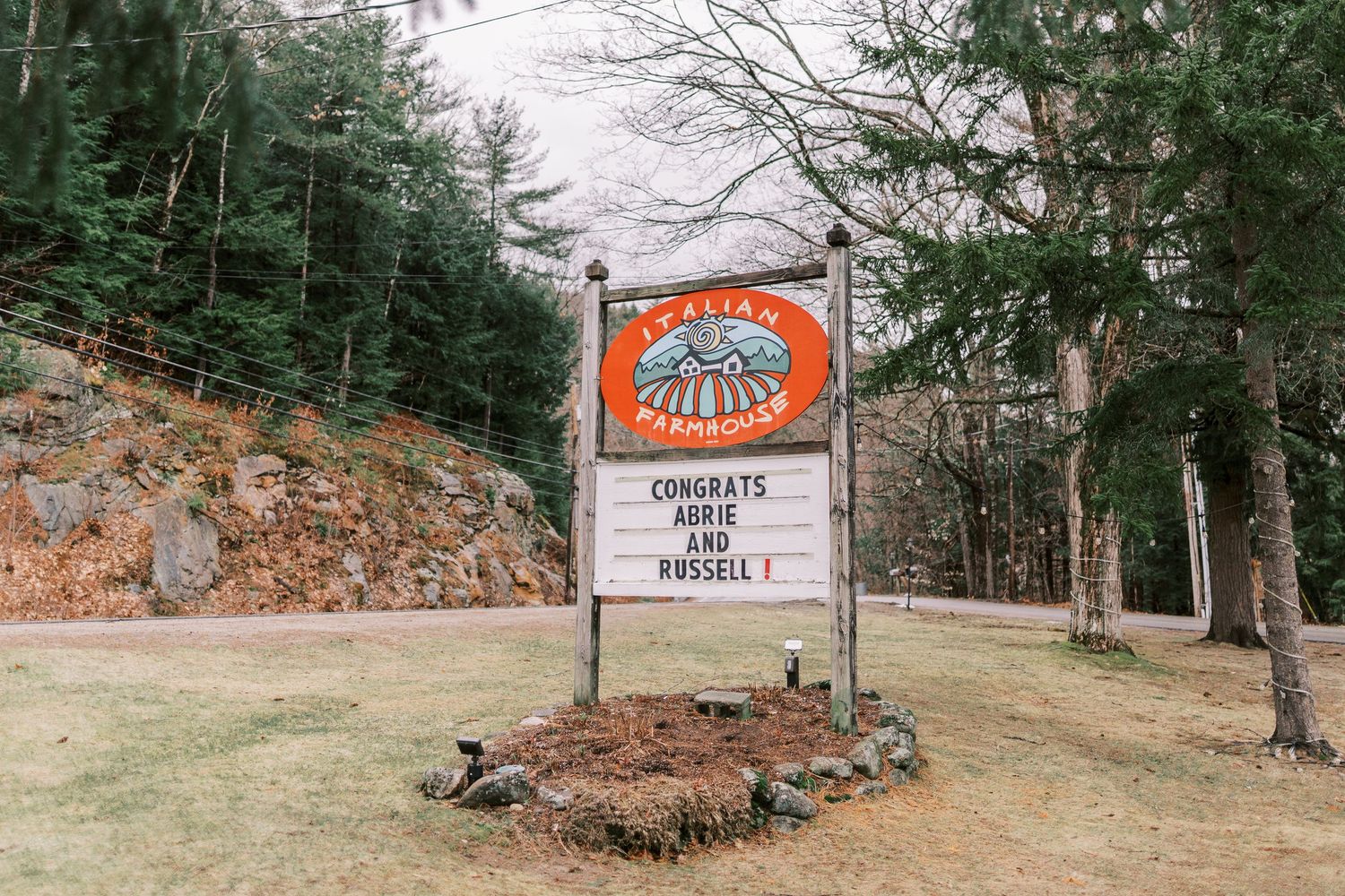 Rustic wooden sign with tribal sun logo among evergreen trees and stone wall at outdoor venue entrance.