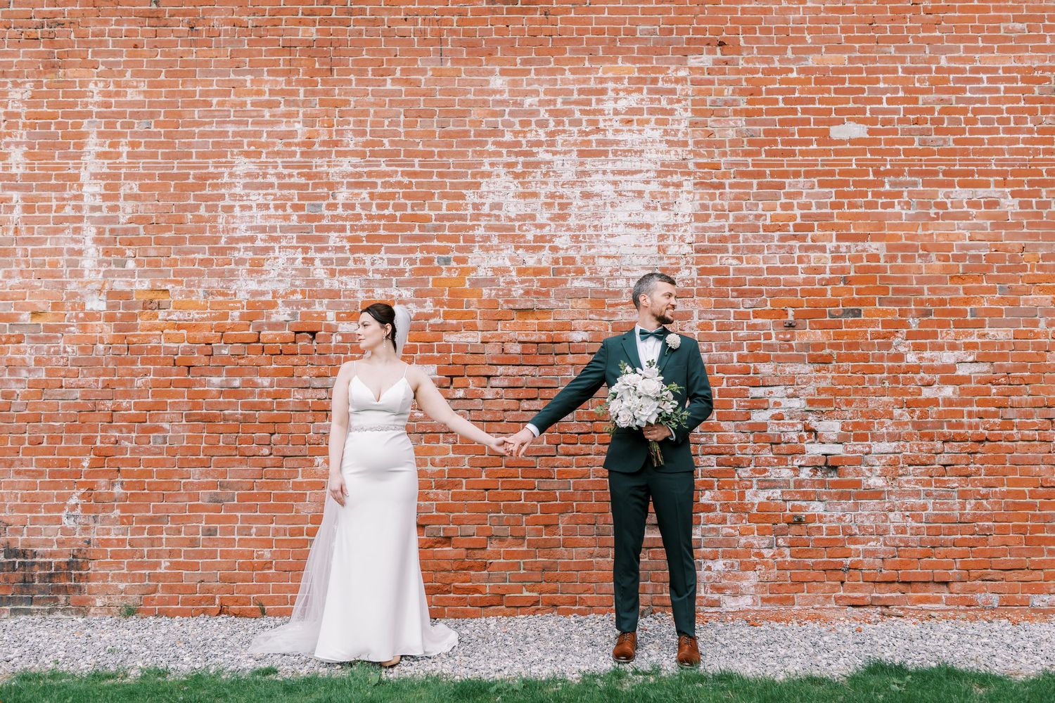 A couple poses creatively against a rustic brick wall during their wedding photoshoot.