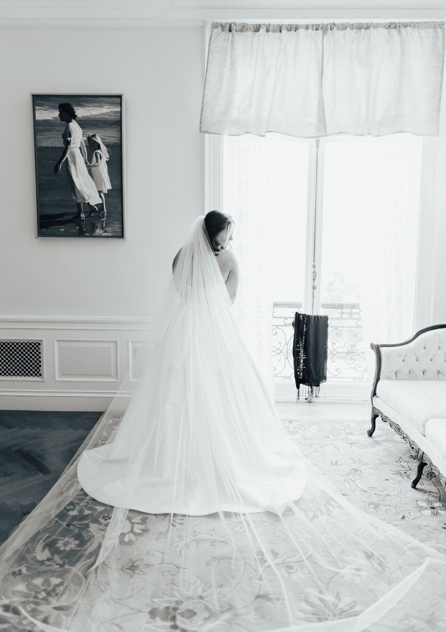 Black and white photograph of a bride in flowing white wedding gown and long veil standing by a bright window.