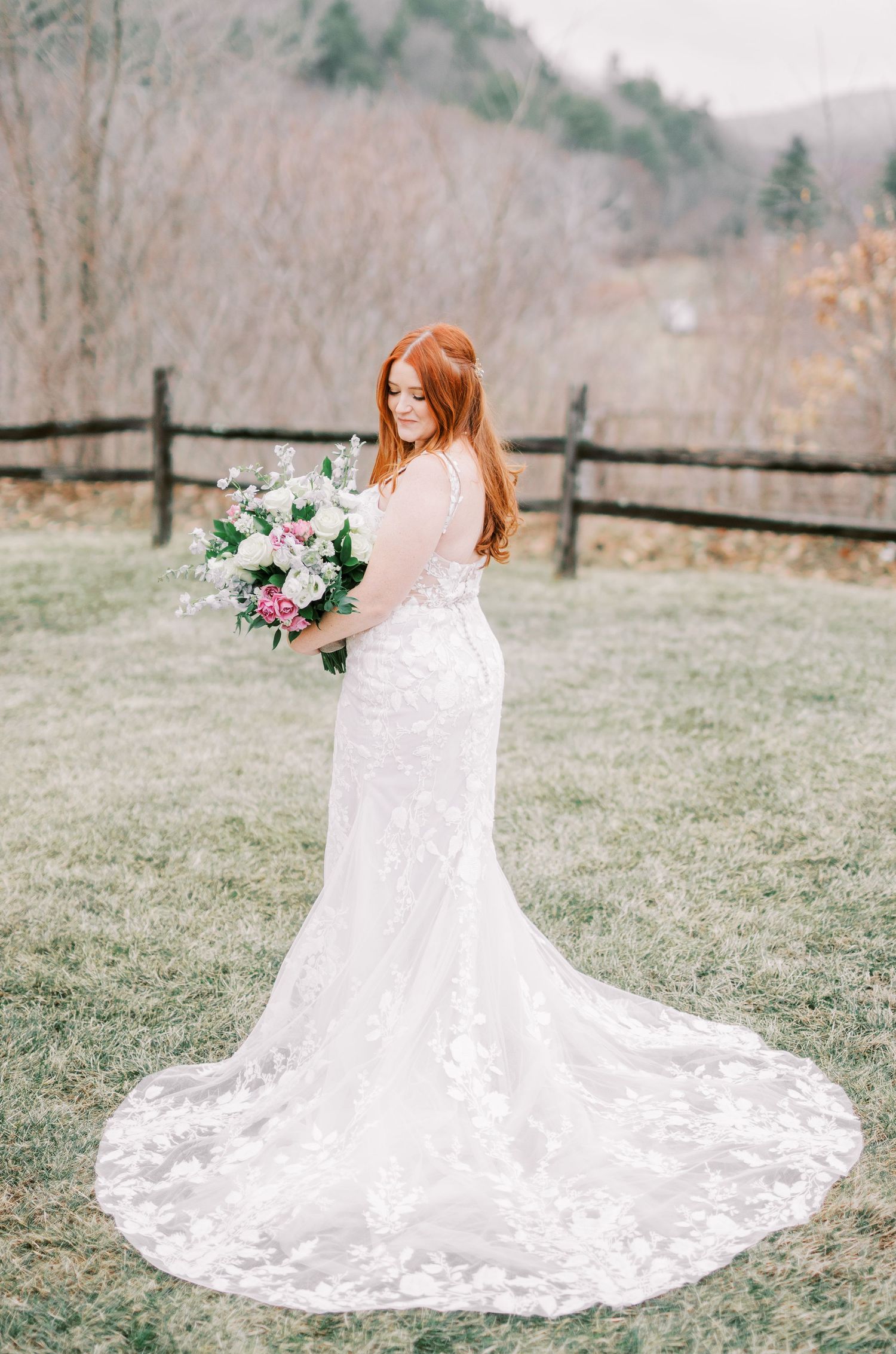 Full-length portrait of bride in lace gown with cathedral train against rustic fence and mountains.