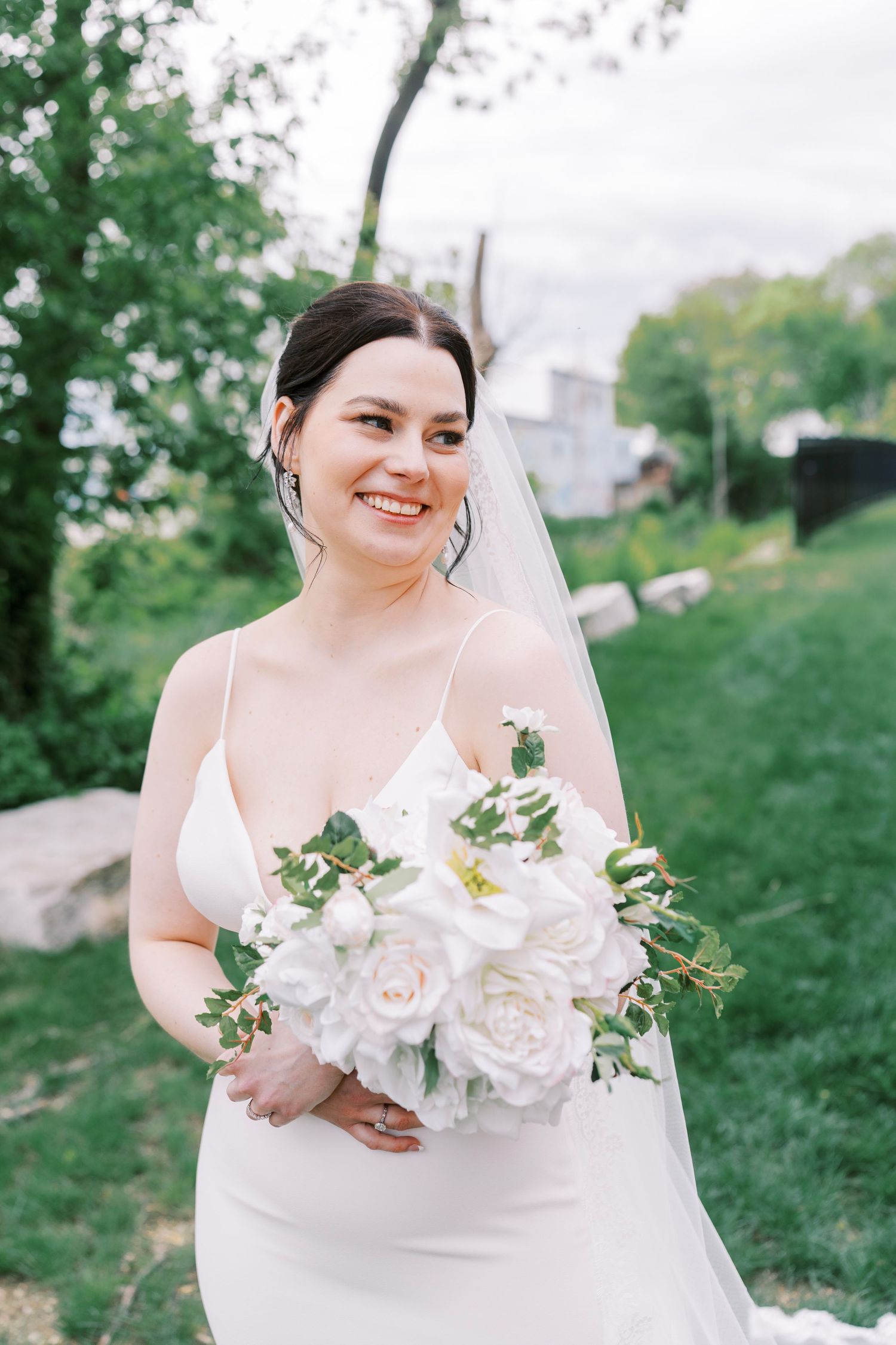 A joyful bride in a white spaghetti strap dress holds a bouquet in an outdoor garden setting.
