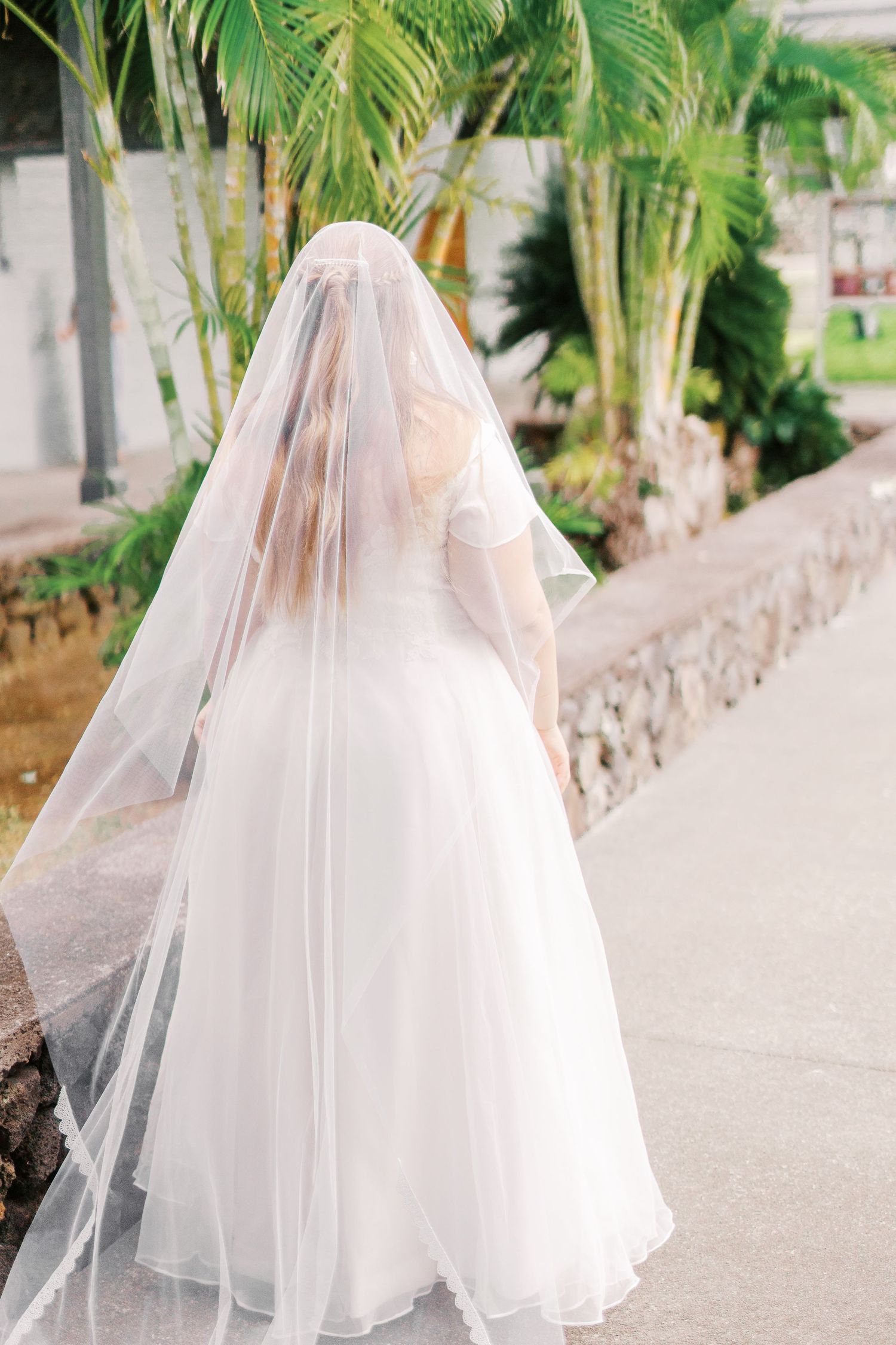 Bride in flowing white dress and veil standing on tropical garden pathway.