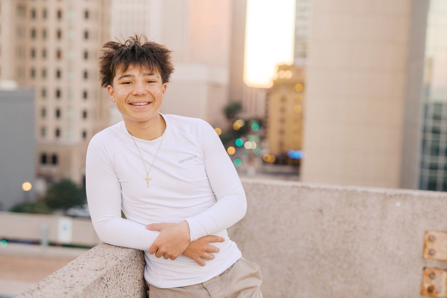 Someone in a white long sleeve shirt smiles while sitting on a ledge with urban buildings in the background.