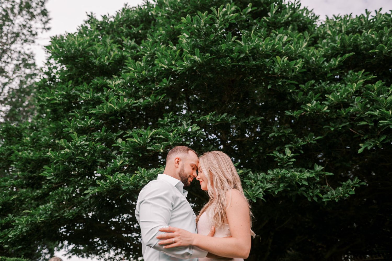 A romantic couple embraces in front of lush green trees during an outdoor photoshoot.