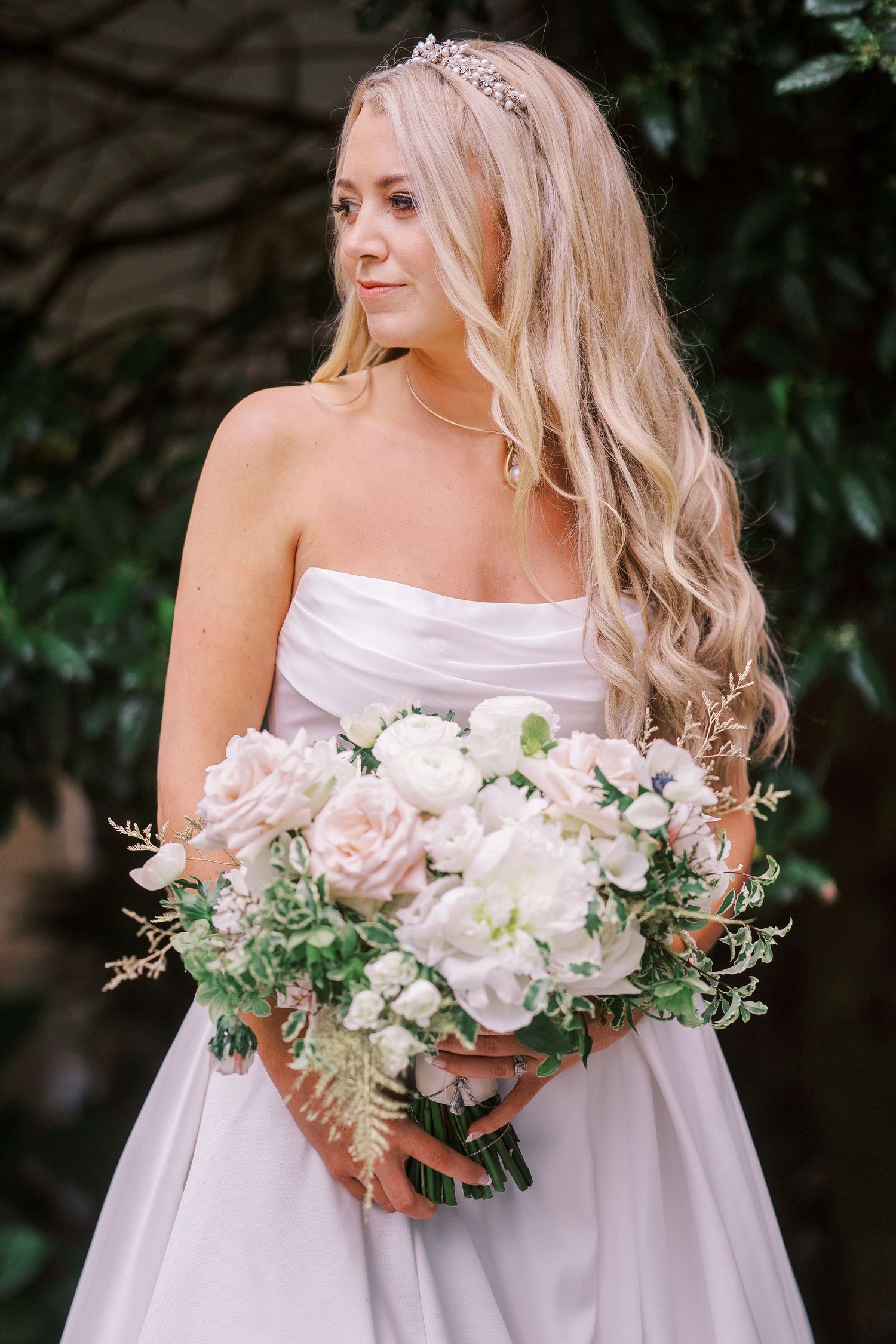 A bride in a strapless white gown holds a romantic bouquet of blush pink roses and white flowers with cascading greenery.