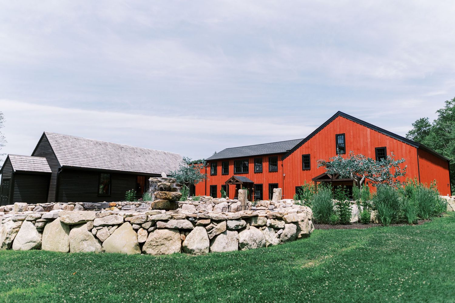 A rustic red barn-style building with a stone retaining wall sits on a grassy landscape under cloudy skies.