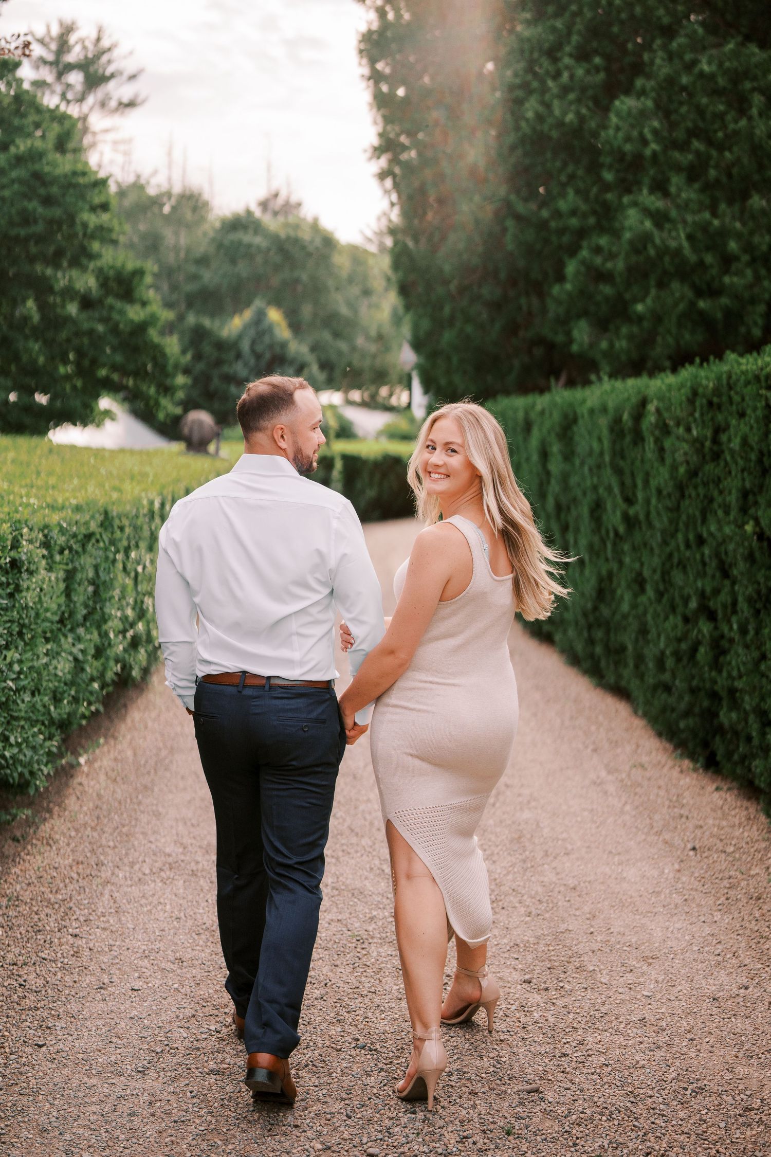 Couple walks playfully down garden path lined with manicured hedges.