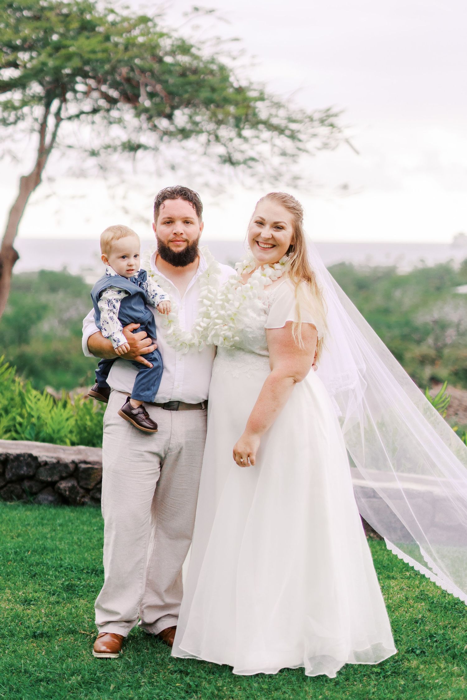 A couple in wedding attire poses outdoors with a small child against a scenic backdrop.