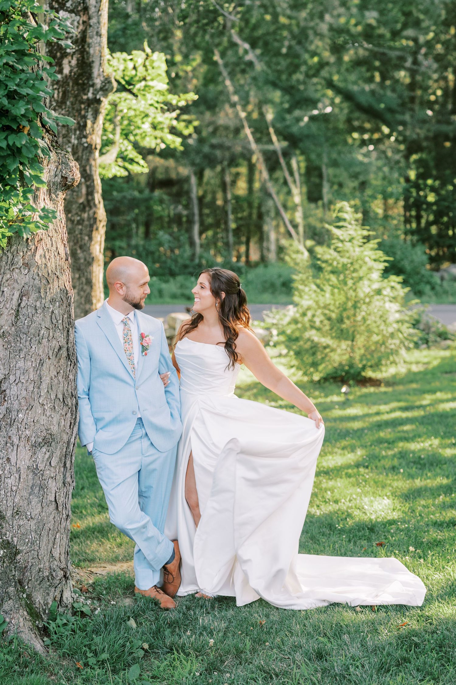 A couple in wedding attire pose playfully by a tree in a sunlit forest setting.