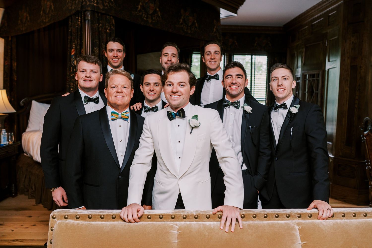 Group of groomsmen in formal black tuxedos and bow ties gather around man in white tuxedo jacket at fancy venue.