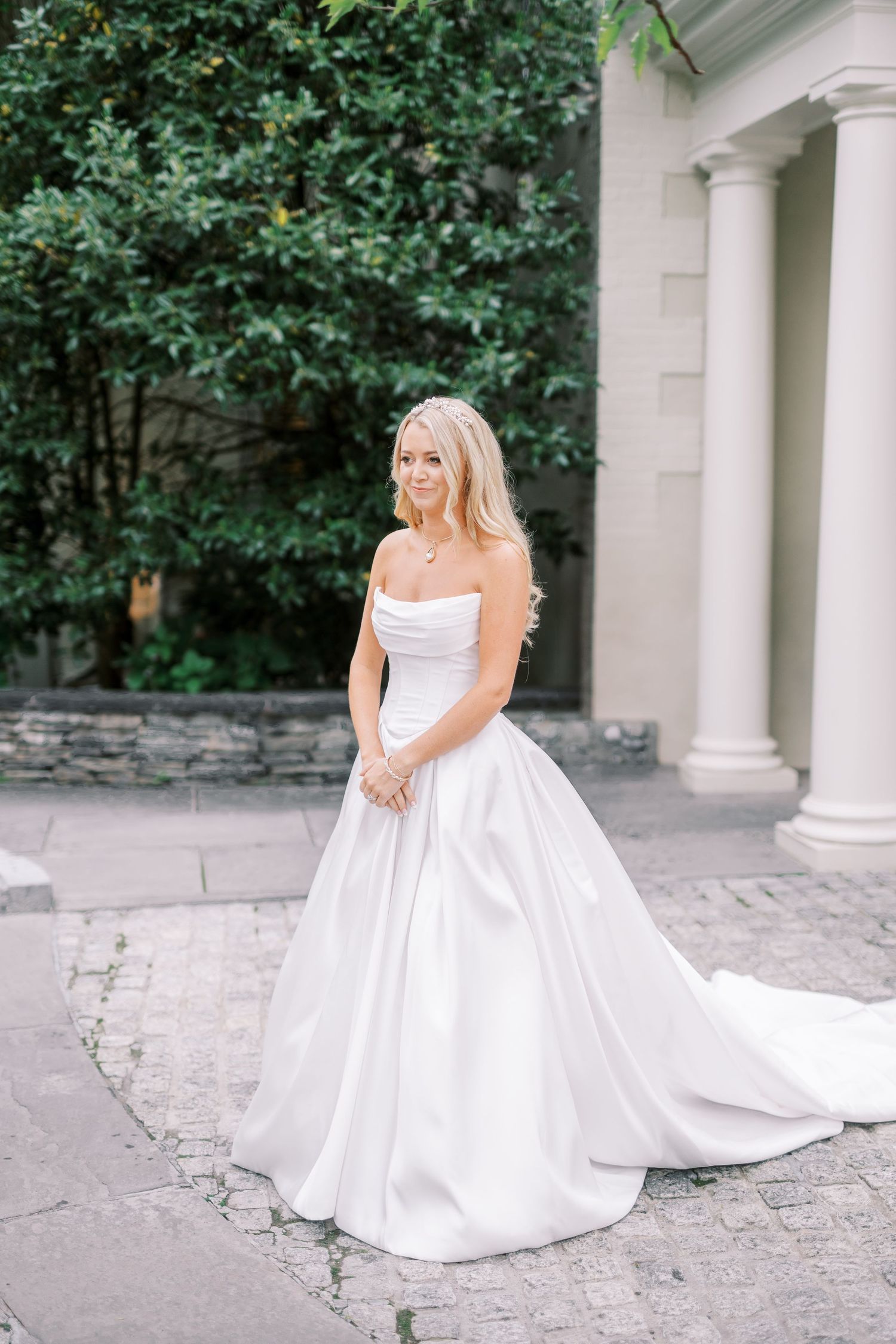 An elegant bride poses on stone pavement in a white strapless ballgown with a long train next to white pillars and greenery.