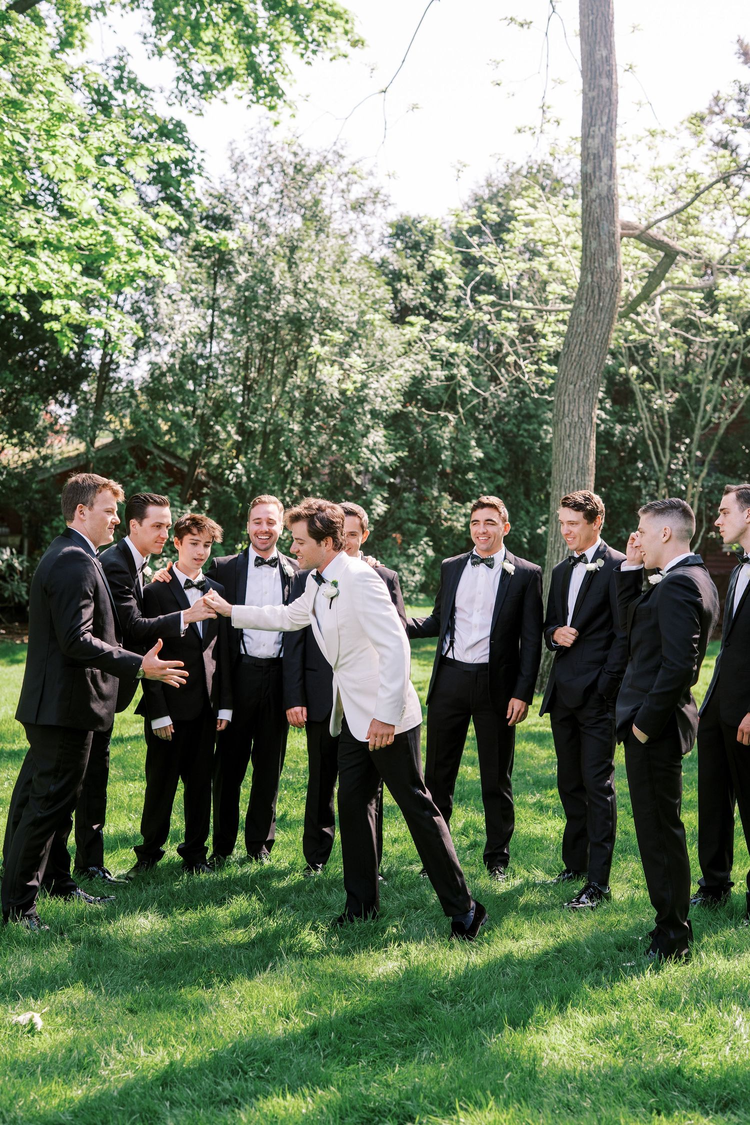 Groomsmen in black tuxedos share a candid moment of laughter on a sunny lawn surrounded by trees.