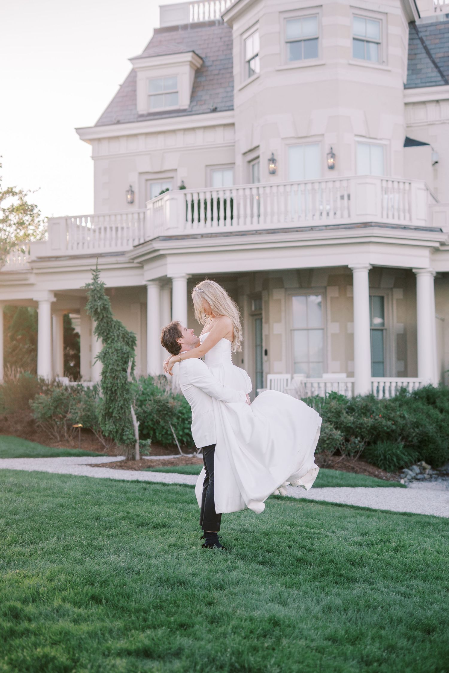 A romantic moment captured outside a grand Victorian mansion with white columns and balcony on a manicured lawn.